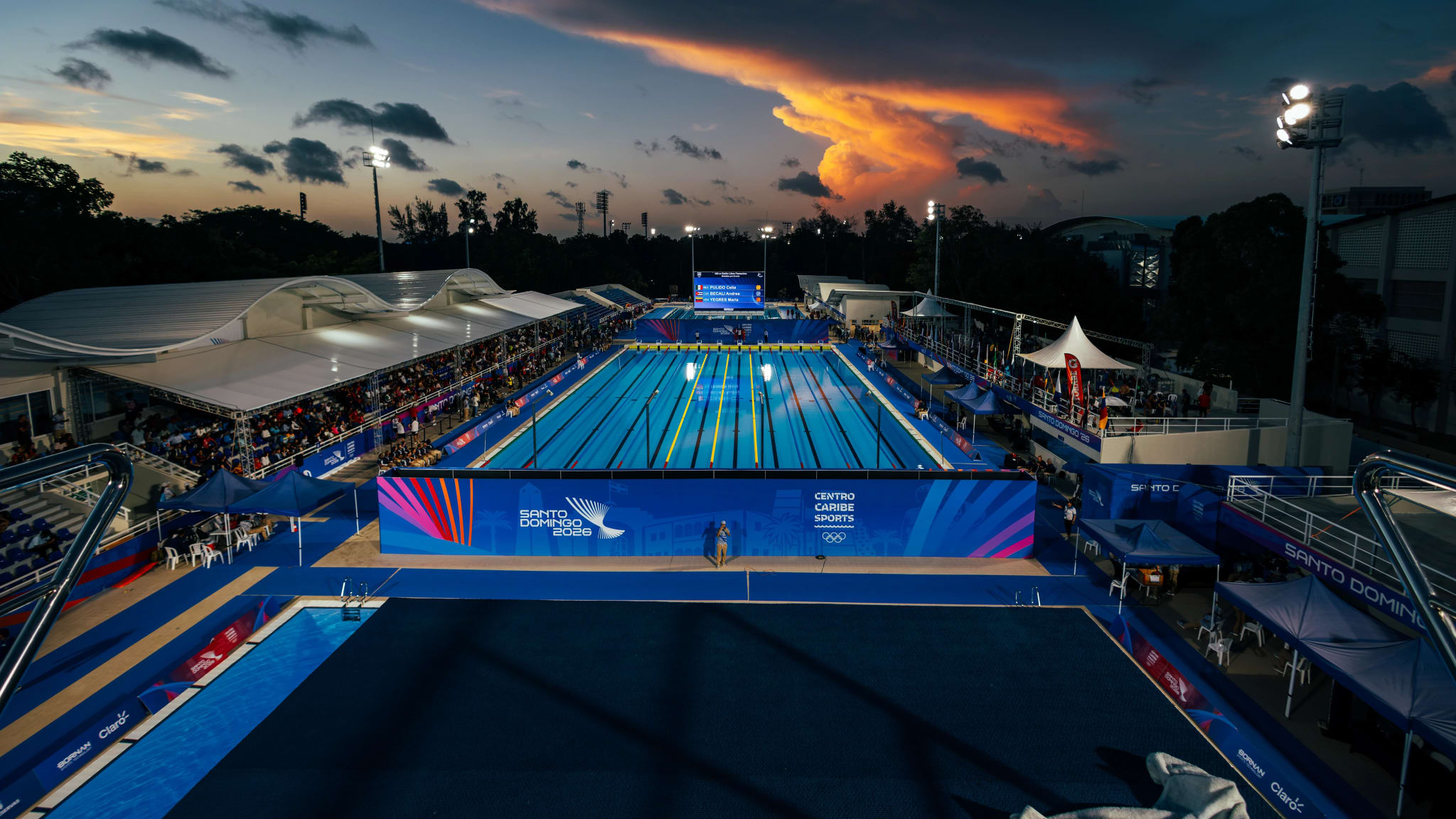 Panoramic view of an outdoor swimming complex at dusk, with dramatic sky and venue lighting