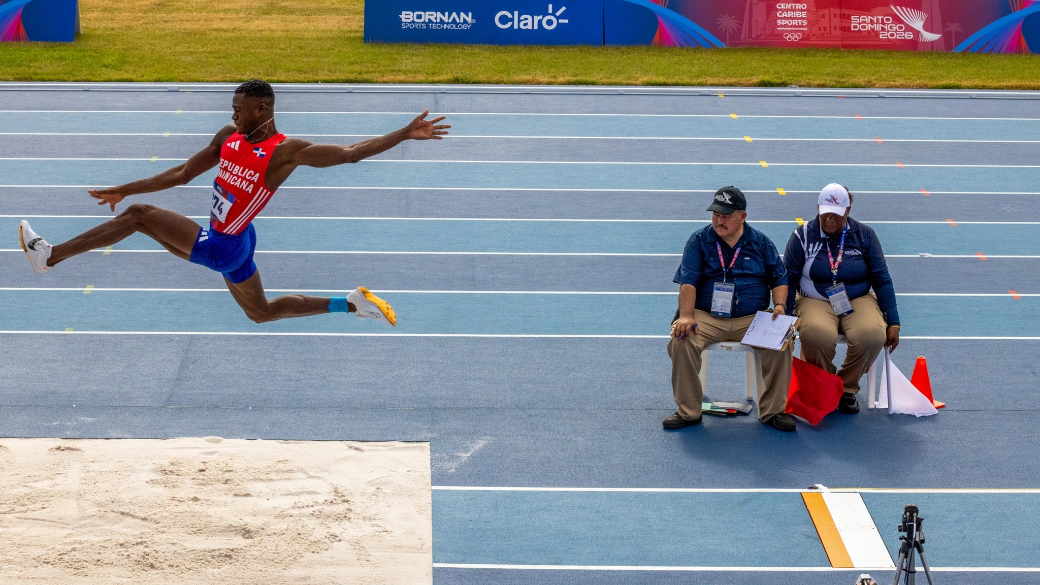Triple jump athlete mid-flight over the track, with judges watching from the sideline
