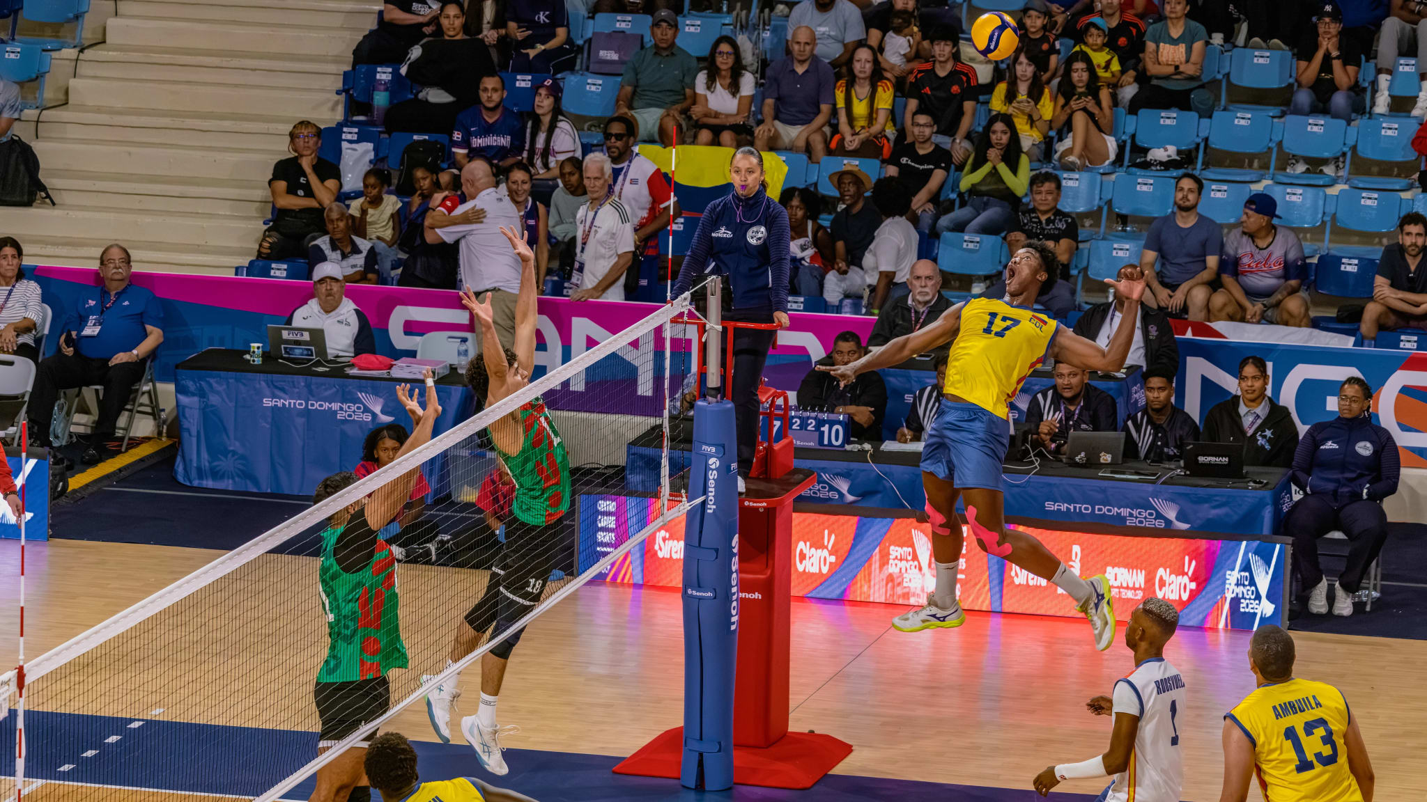 Volleyball hitter jumping to attack over the net, with a rival block and crowd in the stands