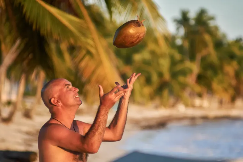 Man on Santo Domingo beach tosses coconut skyward while smiling, palm trees and sea in background, warm golden morning light, family photography session