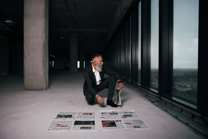 Bearded man in black suit crouching by high-rise window overlooking Santo Domingo, diffuse natural light, professional corporate portrait