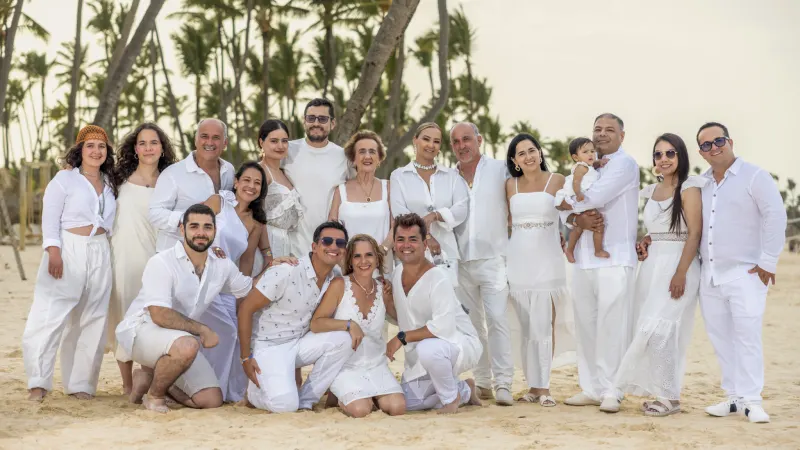Multigenerational family photography session on Punta Cana beach, sixteen people dressed in white under palm trees with filtered natural light.