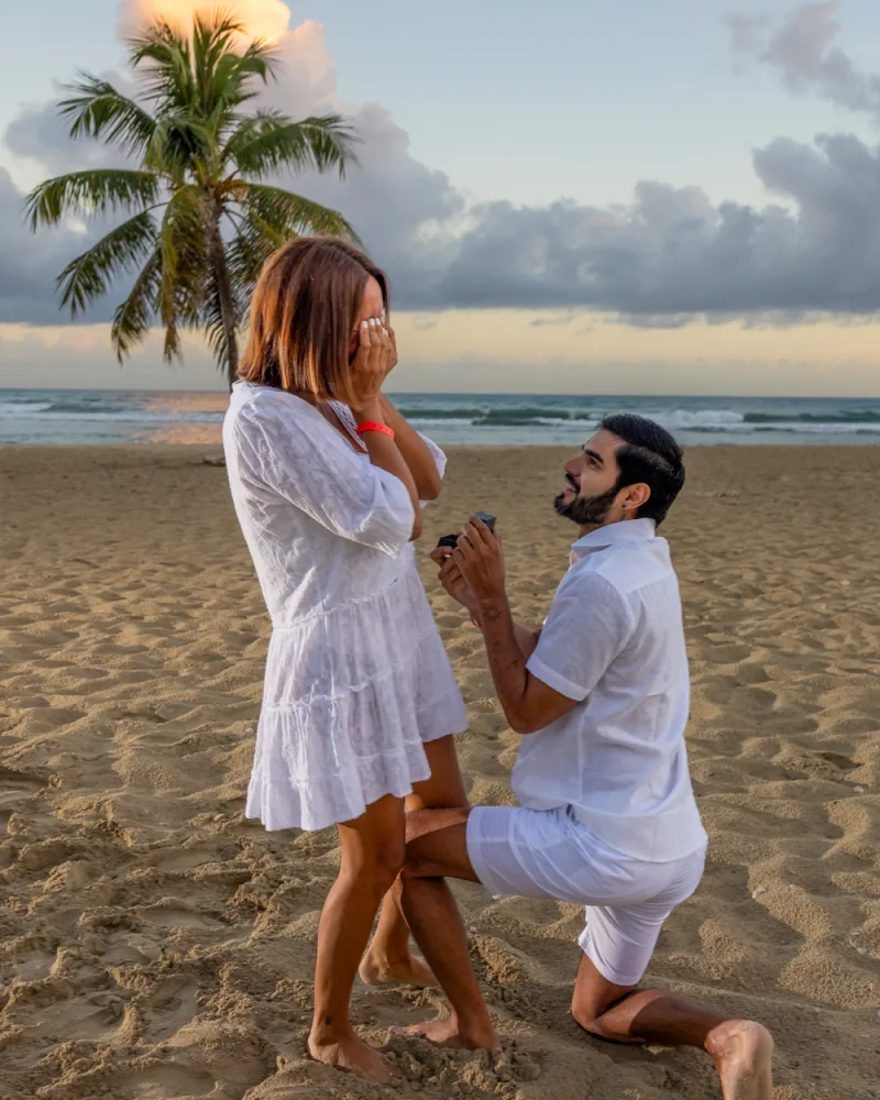 Man kneeling on sand proposes marriage to woman on beach at sunset in Dominican Republic, with palm tree and ocean in background