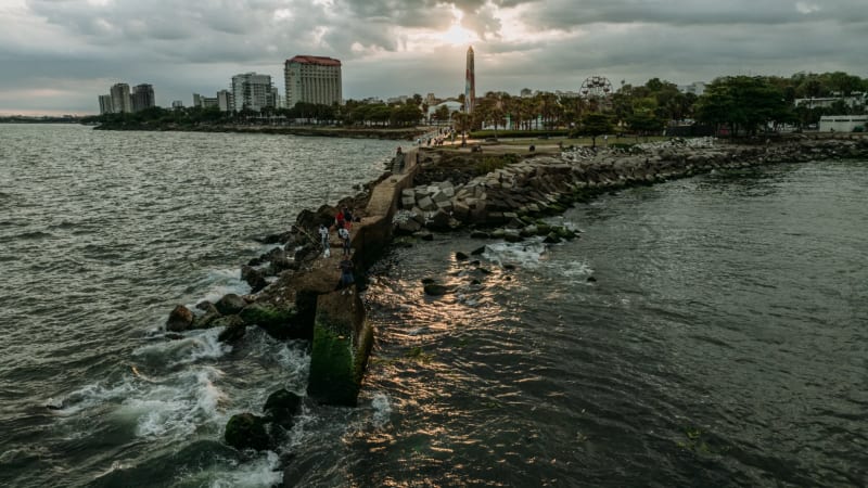 Family posing on the jetty of Santo Domingo's Malecón with coastal architecture and cloudy sky in background, professional family photography session
