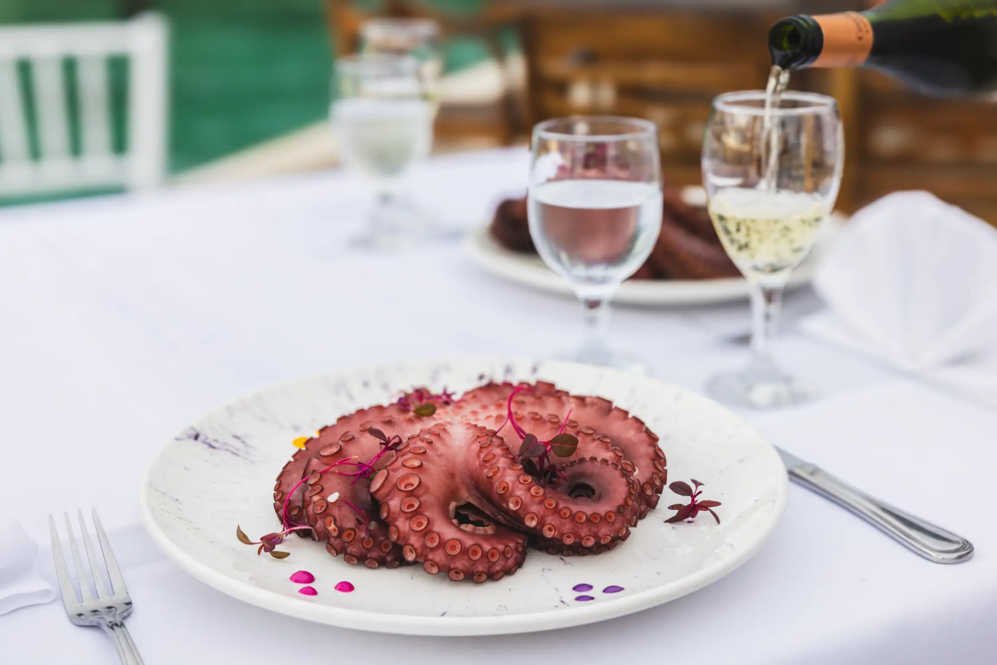 Entrada de pulpo frío decorada con flores comestibles, copa de champán sirviéndose y vista a la marina de fondo