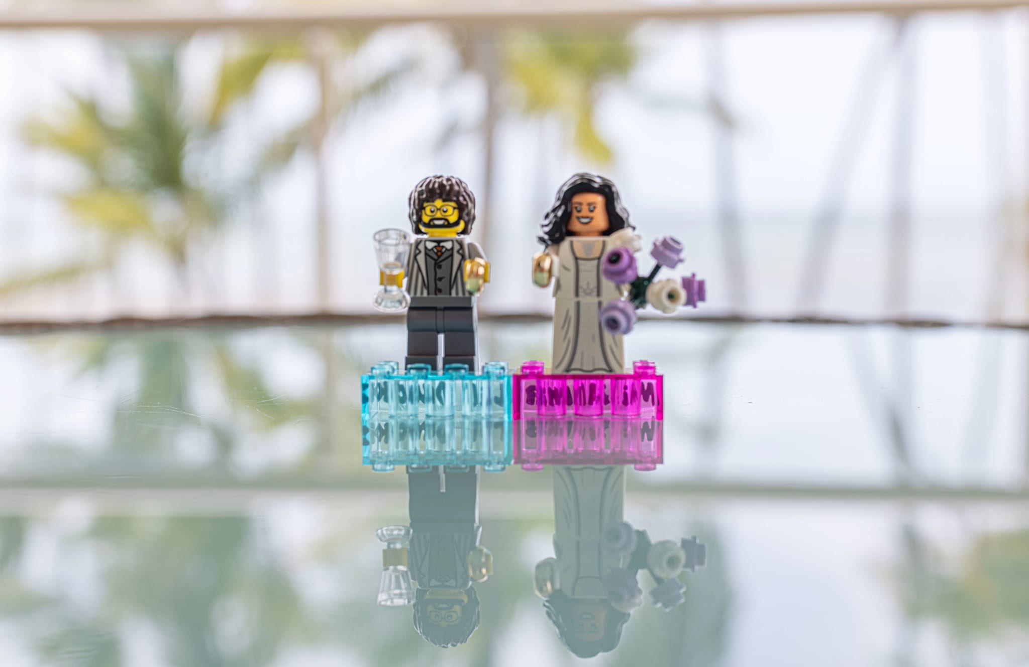 Figuras de Lego de novia y novio sobre una mesa de vidrio con palmeras y el mar de fondo