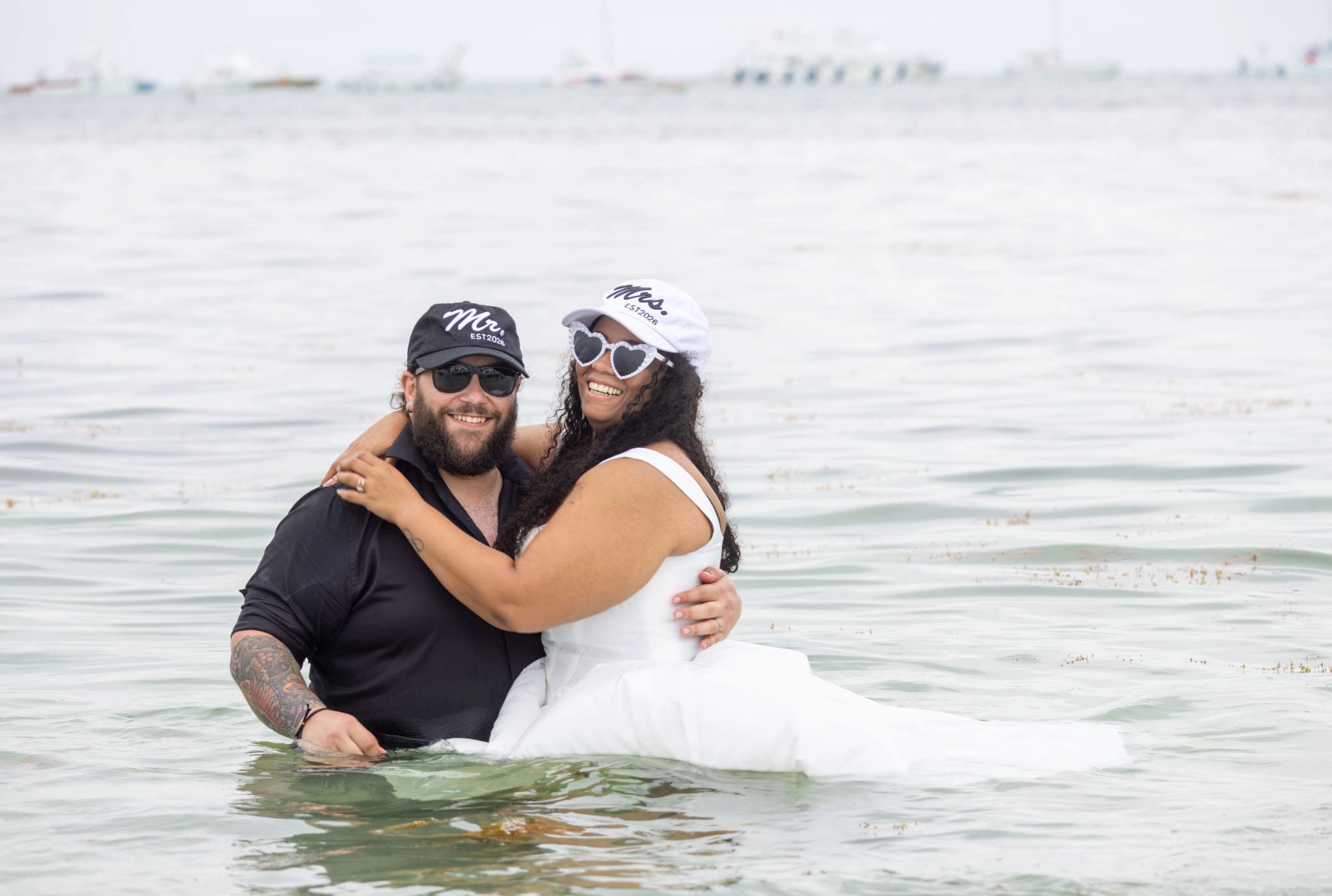 Novios abrazados en el mar durante su sesión de trash the dress, con gorras "Mr." y "Mrs."