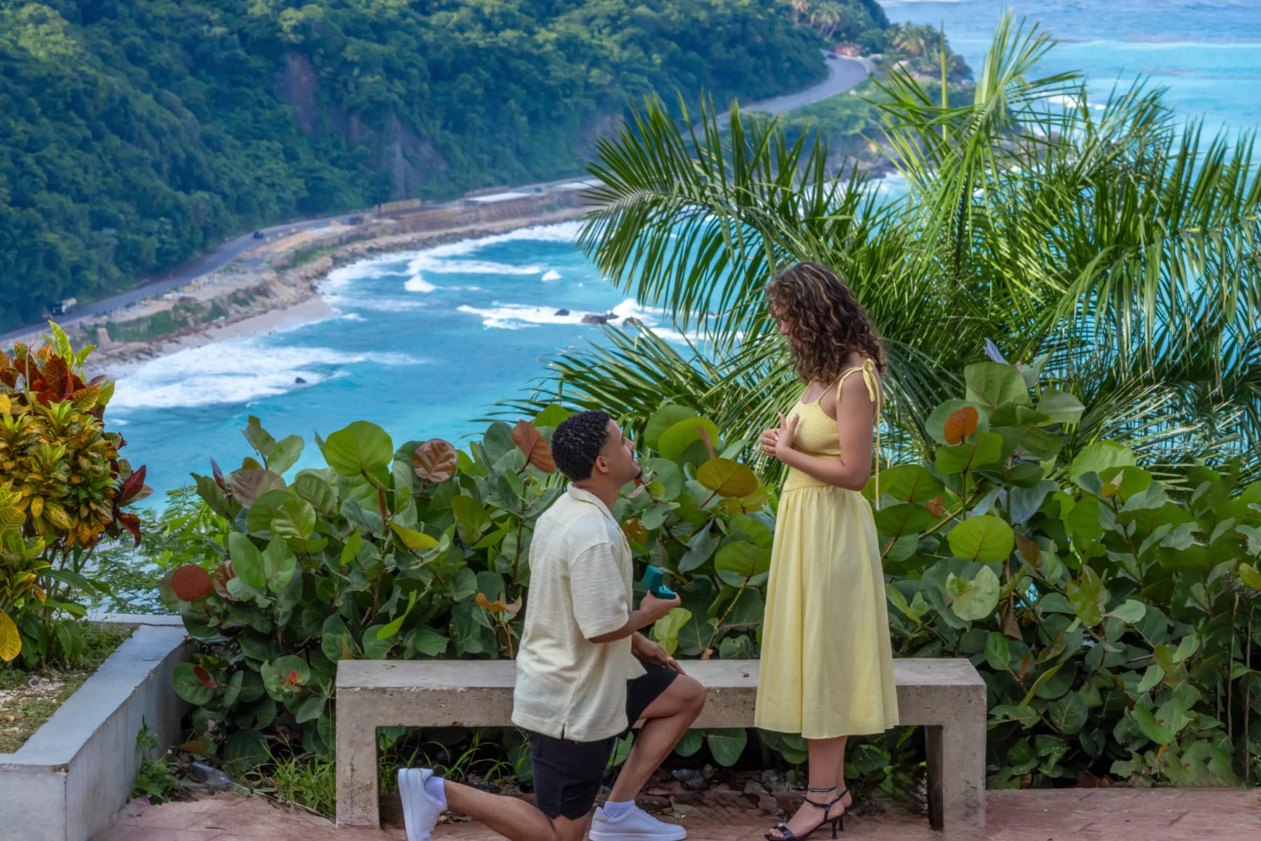 Él se arrodilla para proponerle matrimonio con la caja del anillo abierta, en un mirador sobre el mar Caribe