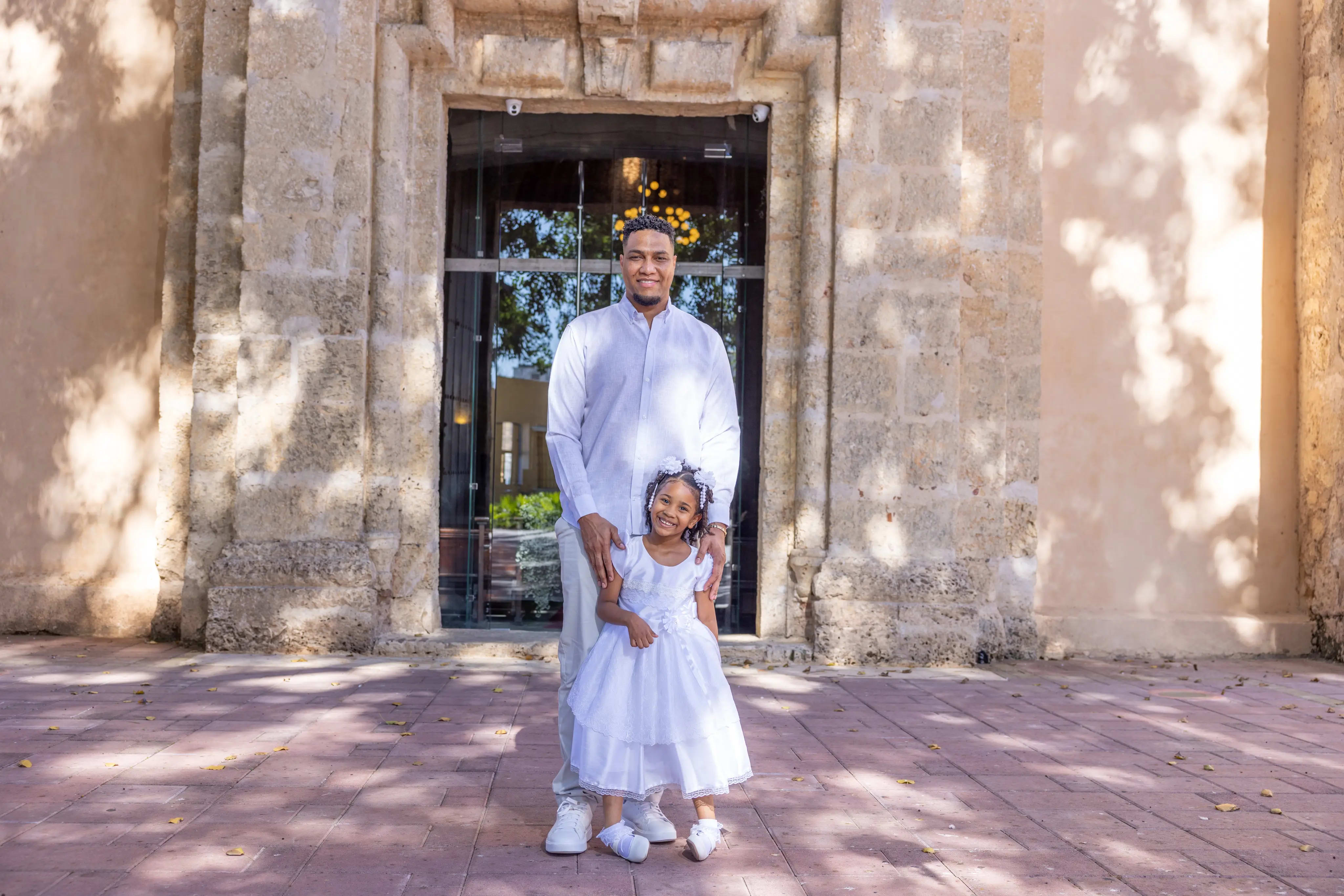Padre e hija con vestido de bautizo blanco llegando a la puerta de piedra de una iglesia histórica en la Zona Colonial, Santo Domingo — Babula Shots