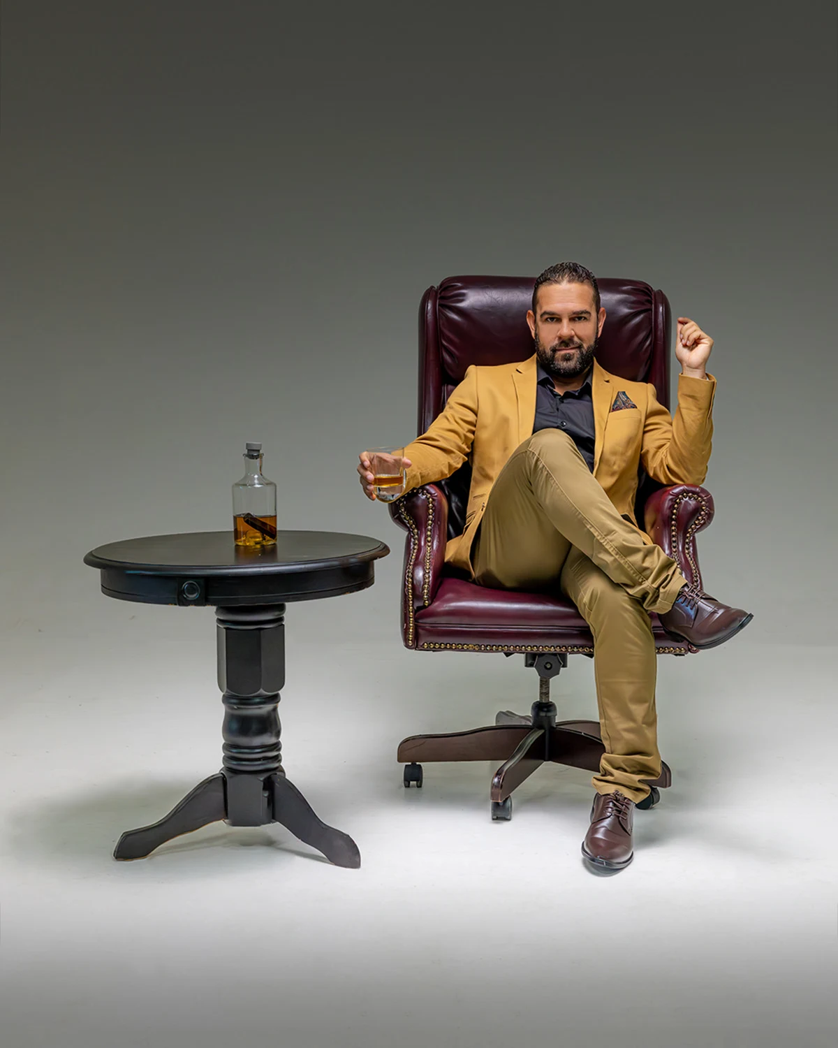 Retrato vertical de estudio de un hombre con saco mostaza y pantalón caqui, sentado en un sillón de cuero burdeos con una copa de whisky en mano