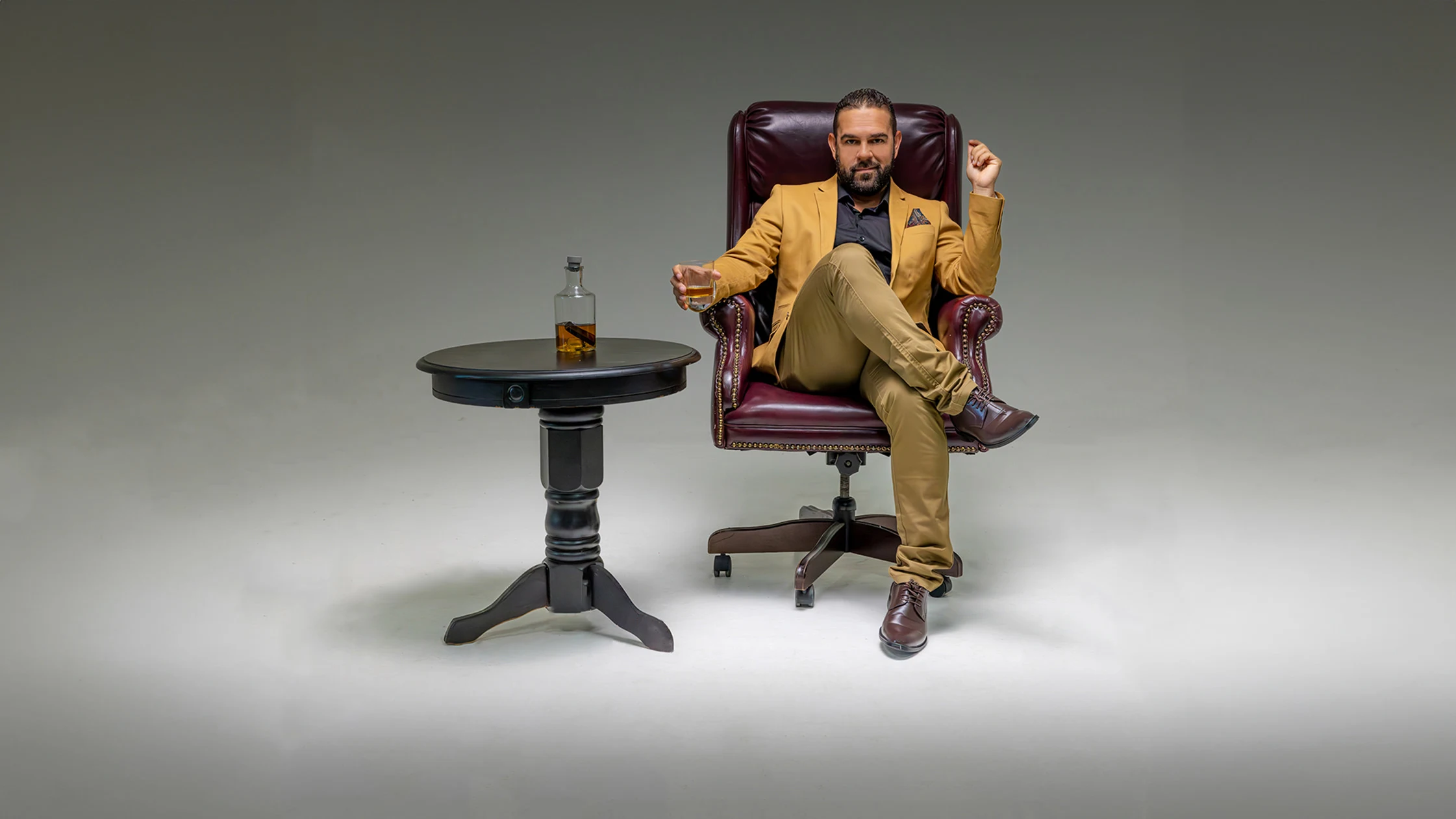 Retrato de estudio de un hombre con saco mostaza sentado en un sillón de cuero color burdeos, sosteniendo una copa de whisky junto a una mesa con decantador
