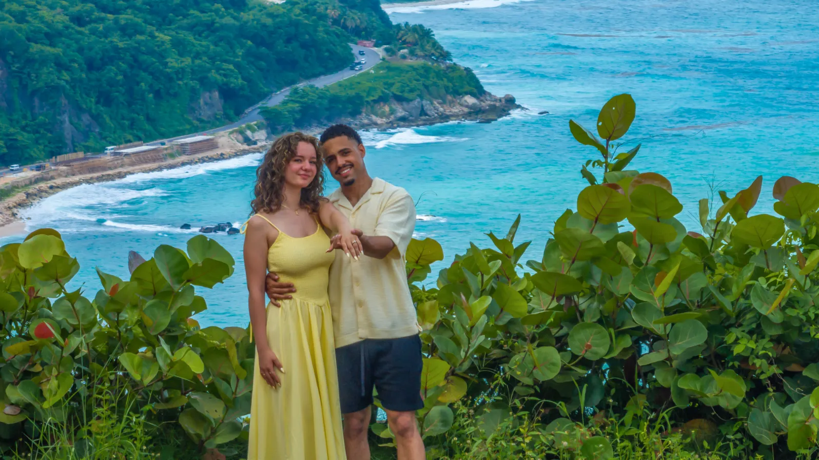 Retrato de compromiso con la pareja sonriendo y extendiendo la mano para mostrar el anillo, con el mirador costero de fondo