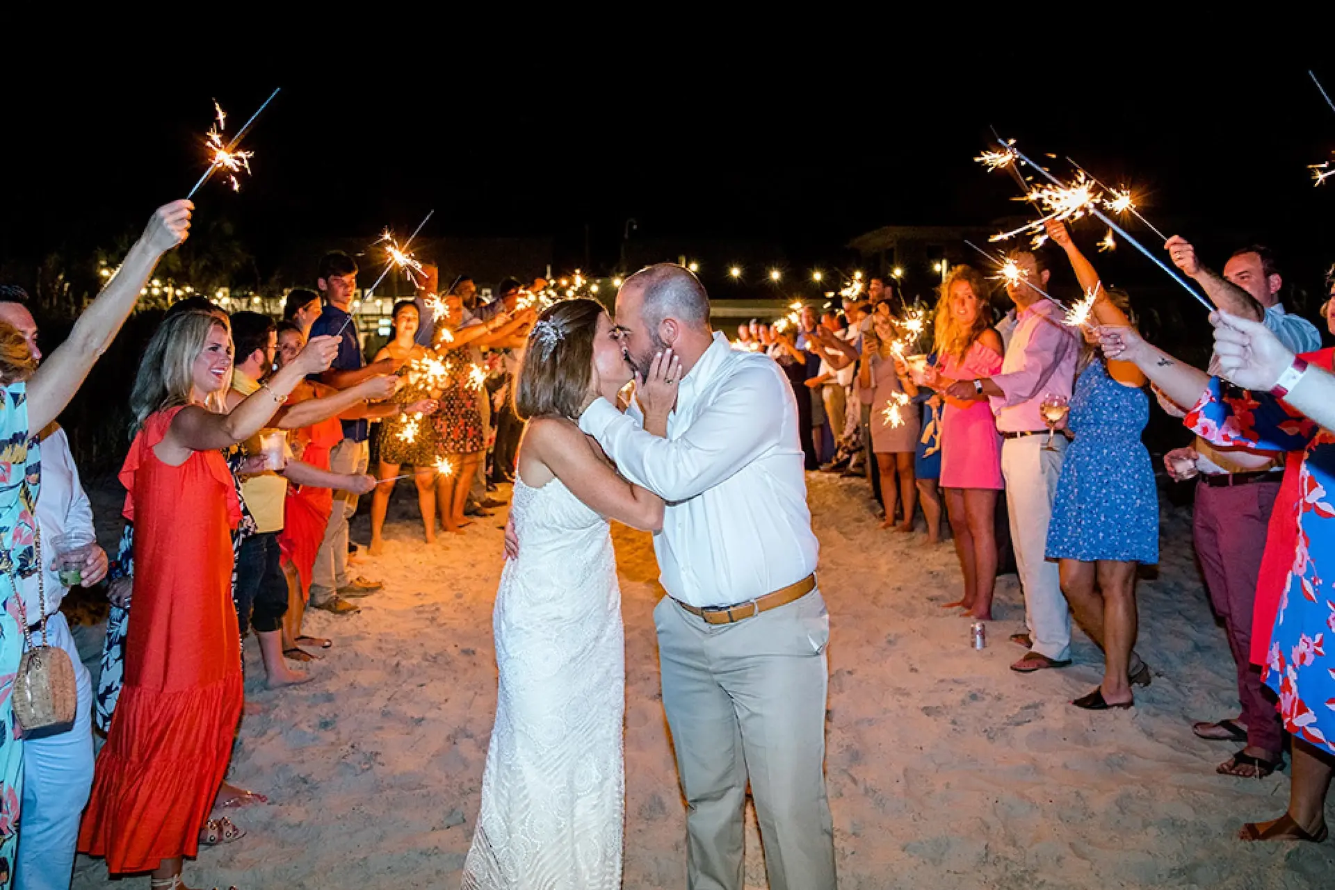 Newlyweds kissing on the beach at night, surrounded by guests with lit sparklers.