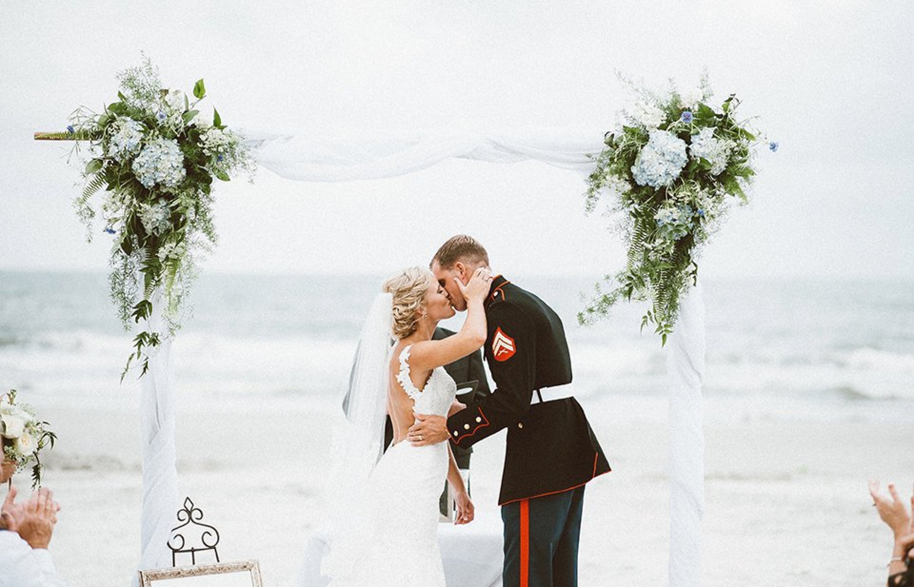 bride and groom in military attire kiss under a wooden arbor with flowers and white tapestry and the ocean as the backdrop