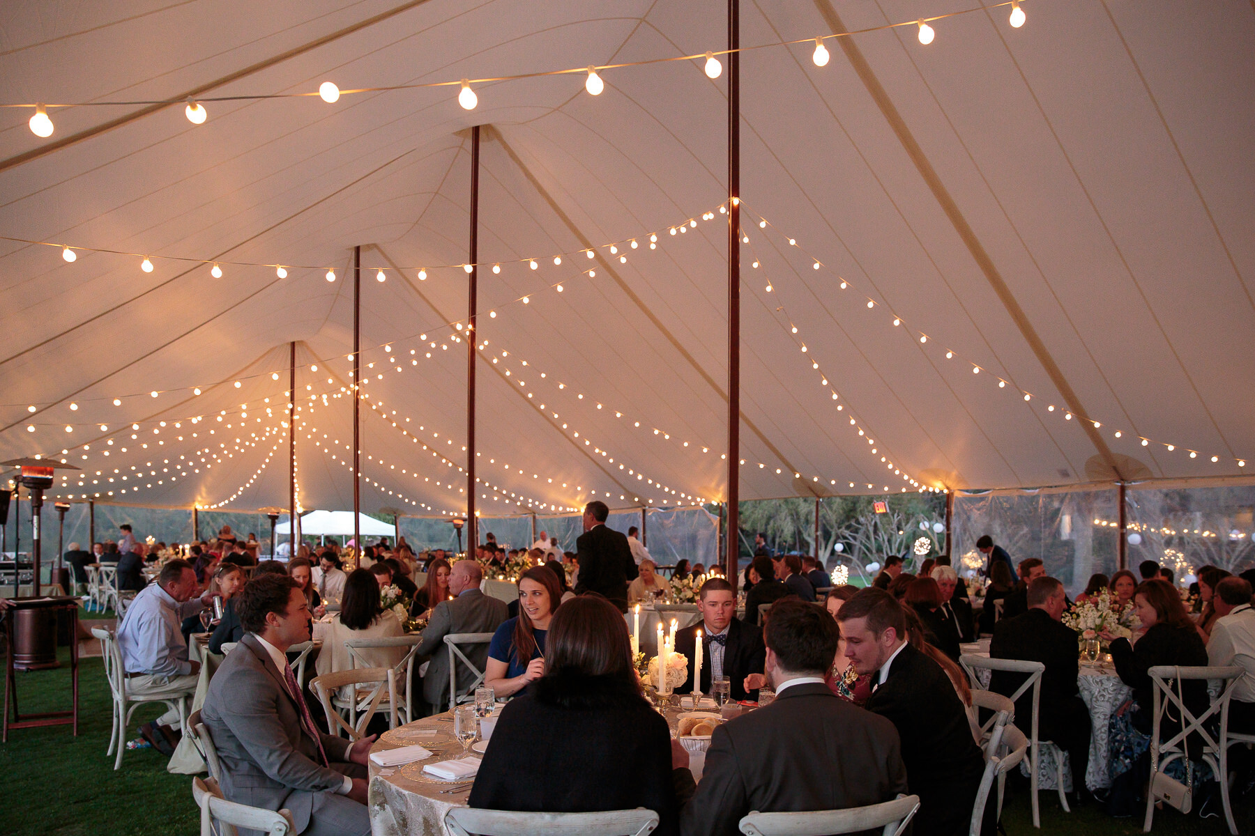 wedding guests sit at their tables under string lights and a large white tent