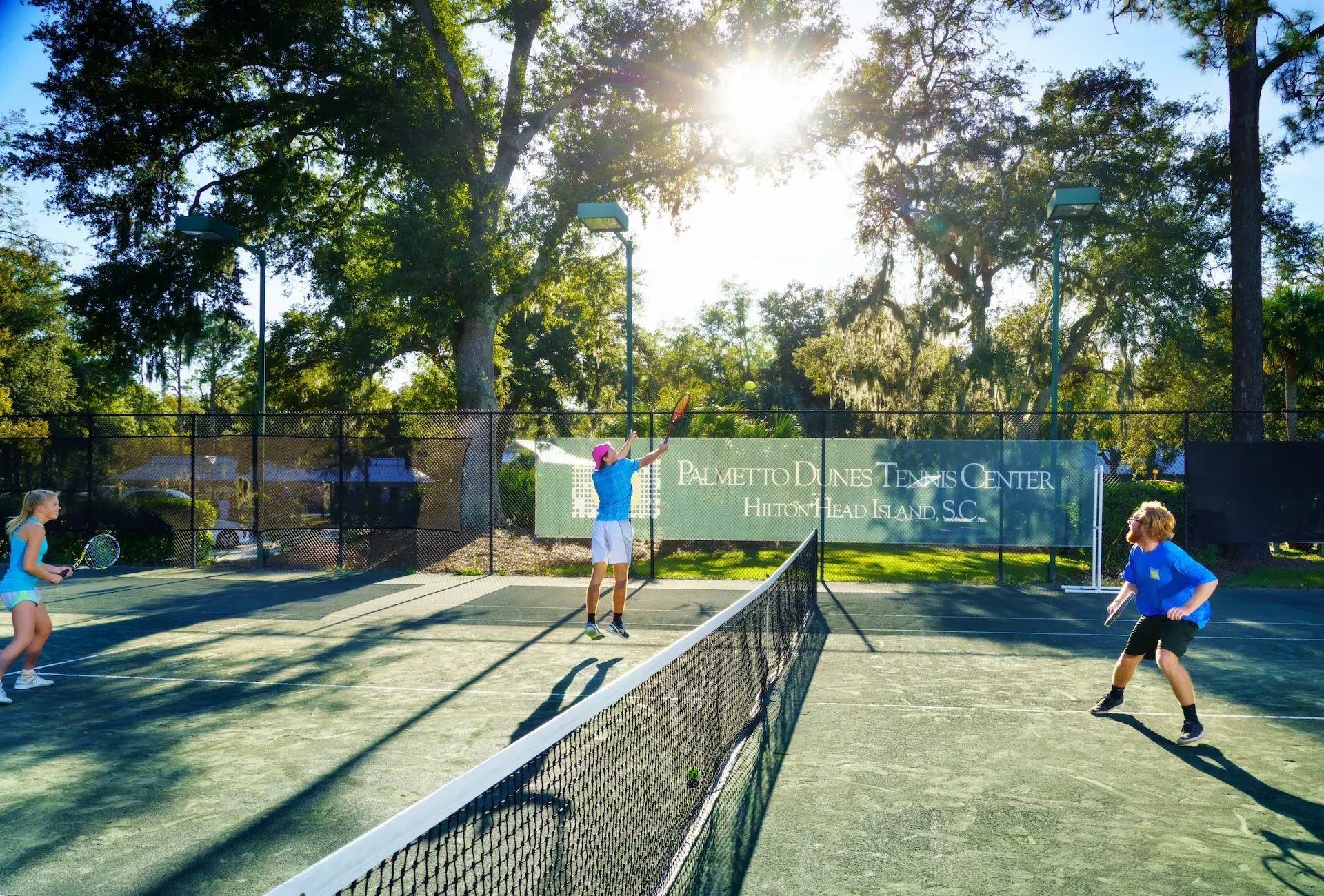 two men actively playing tennis at Palmetto Dunes Tennis center with the sun shining through the trees