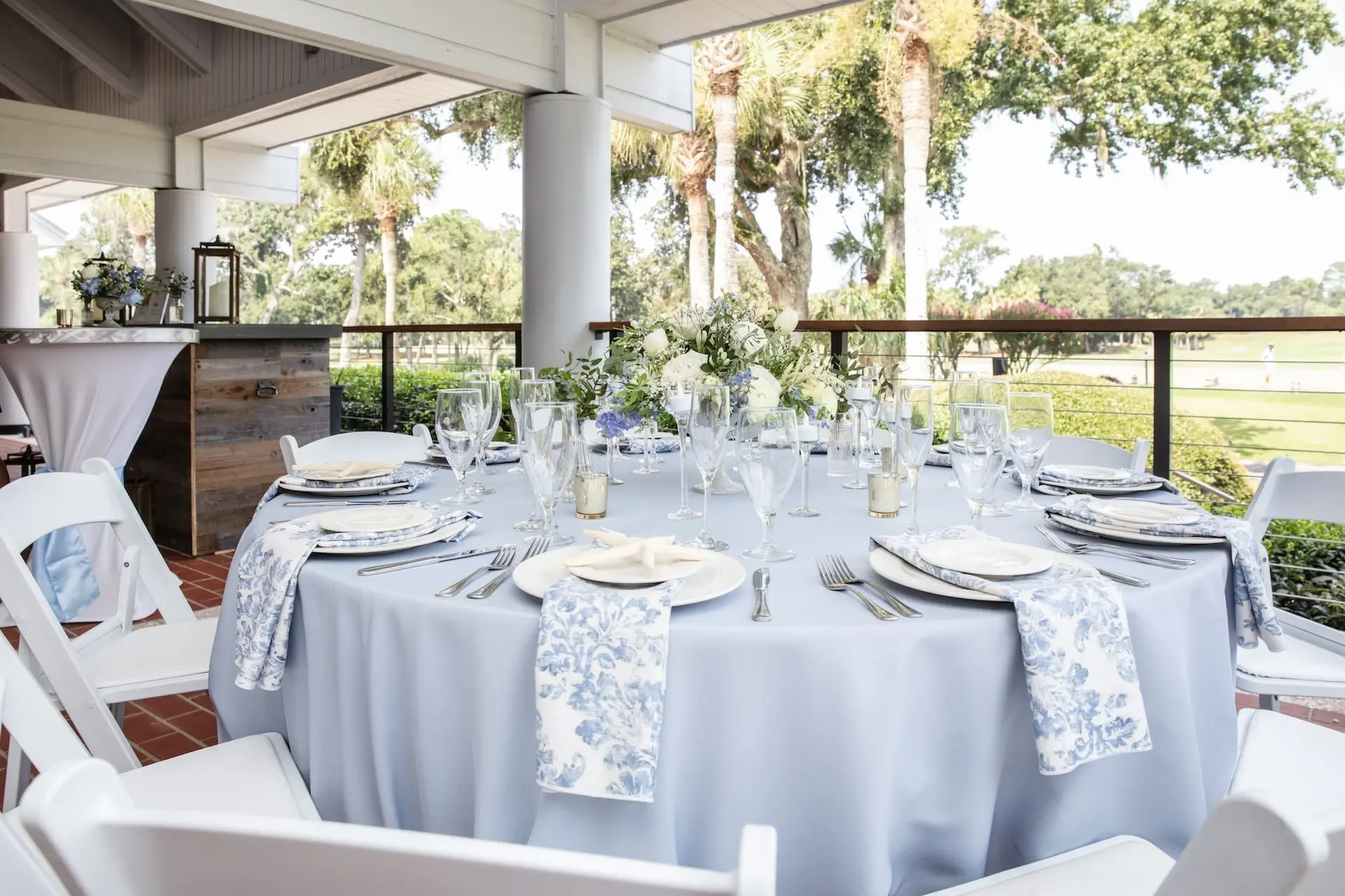Reception table on a terrace, with light blue tablecloths, patterned napkins, and a white flower centerpiece.