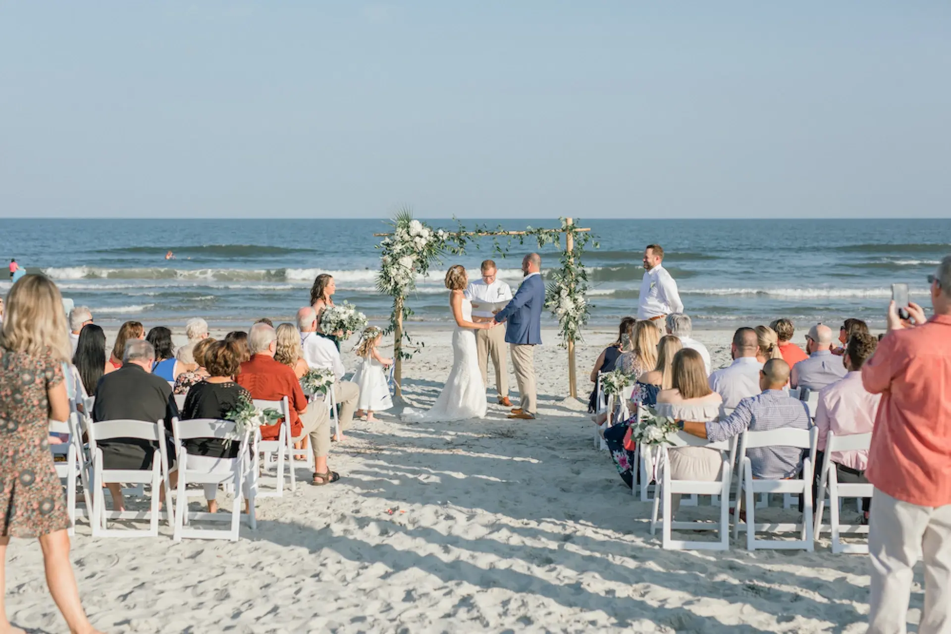 Beach wedding with the couple in front of the flower-decorated wooden altar and guests seated in white chairs.