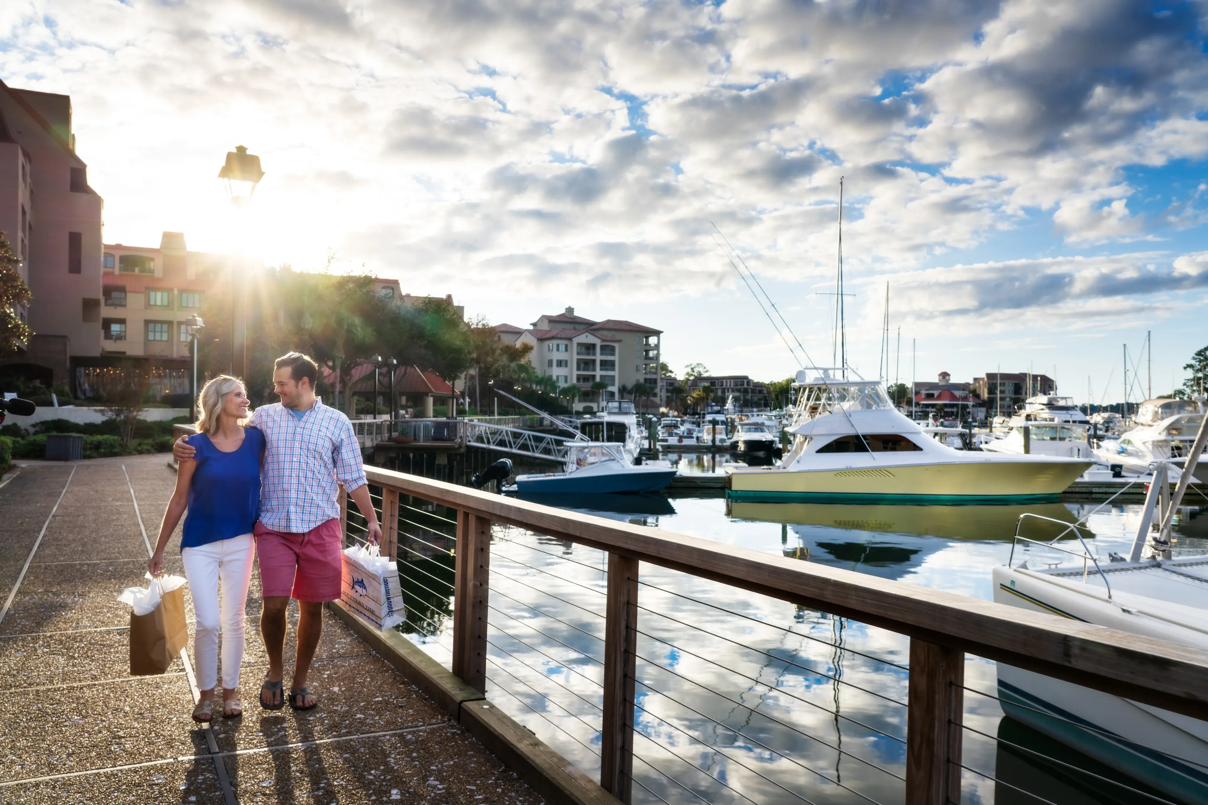 couple embrace each other while holding shopping bags and walk along the promenade next to the marina with the sun shining in the background