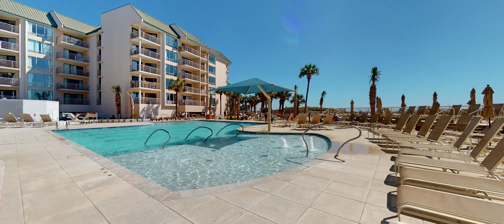 resort style pool surrounded by lounge chairs and a condo building in the background under blue skies