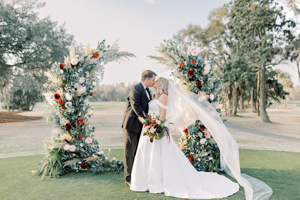 bride and groom kiss under a gorgeous flower arch with trees and greenery in the background