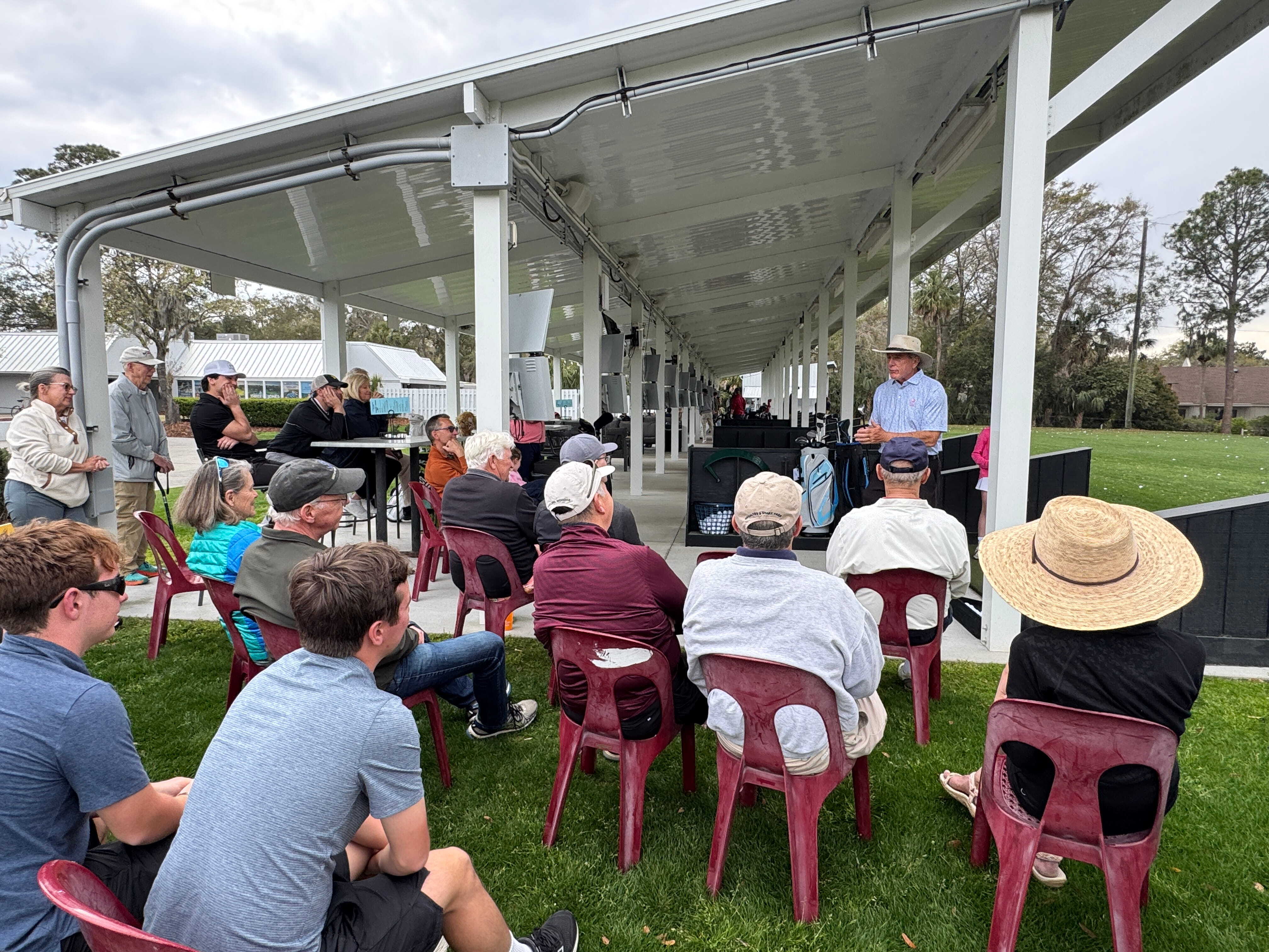 group of spectators listen to Doug Weaver in chairs at the Toptracer Range during a Weekly Clinic