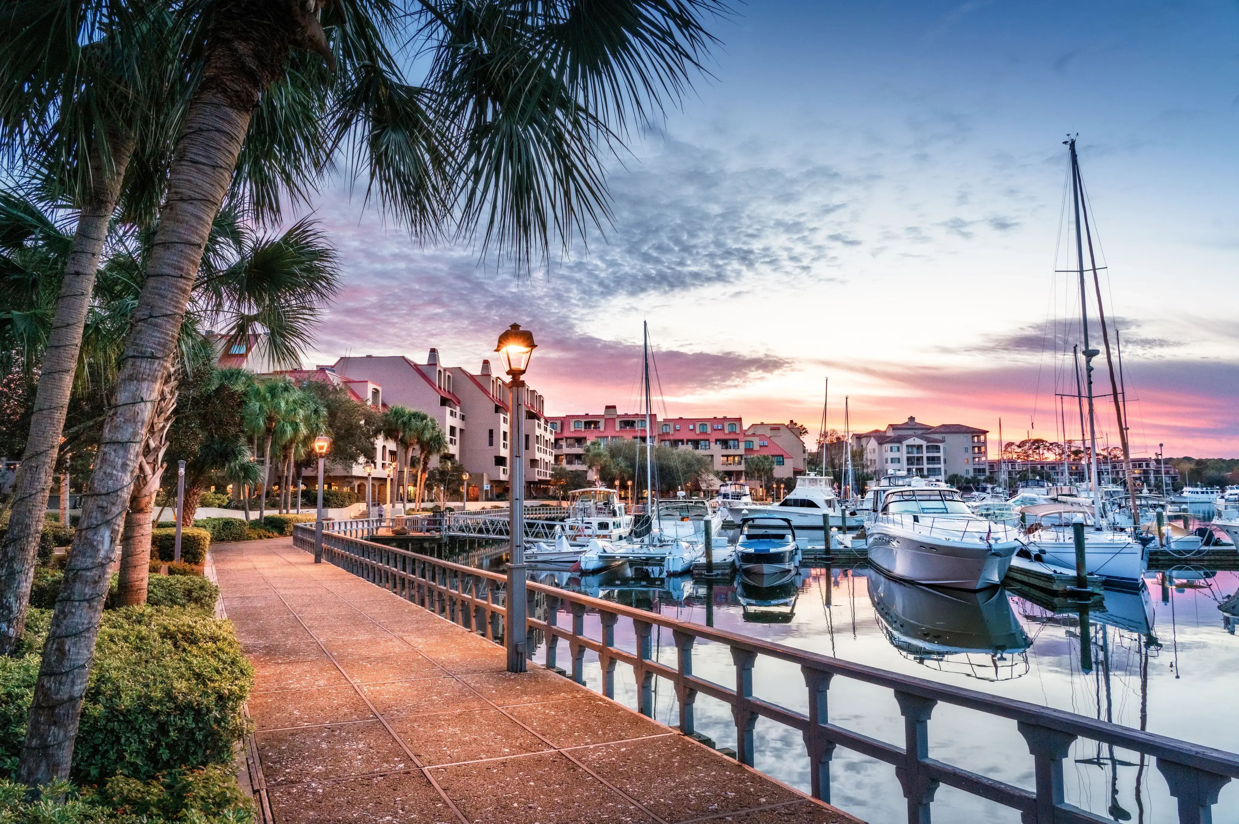 waterfront walkway with the marina and boats on one side and palm trees on the other and the sunsetting in the background painting the sky blue and pink