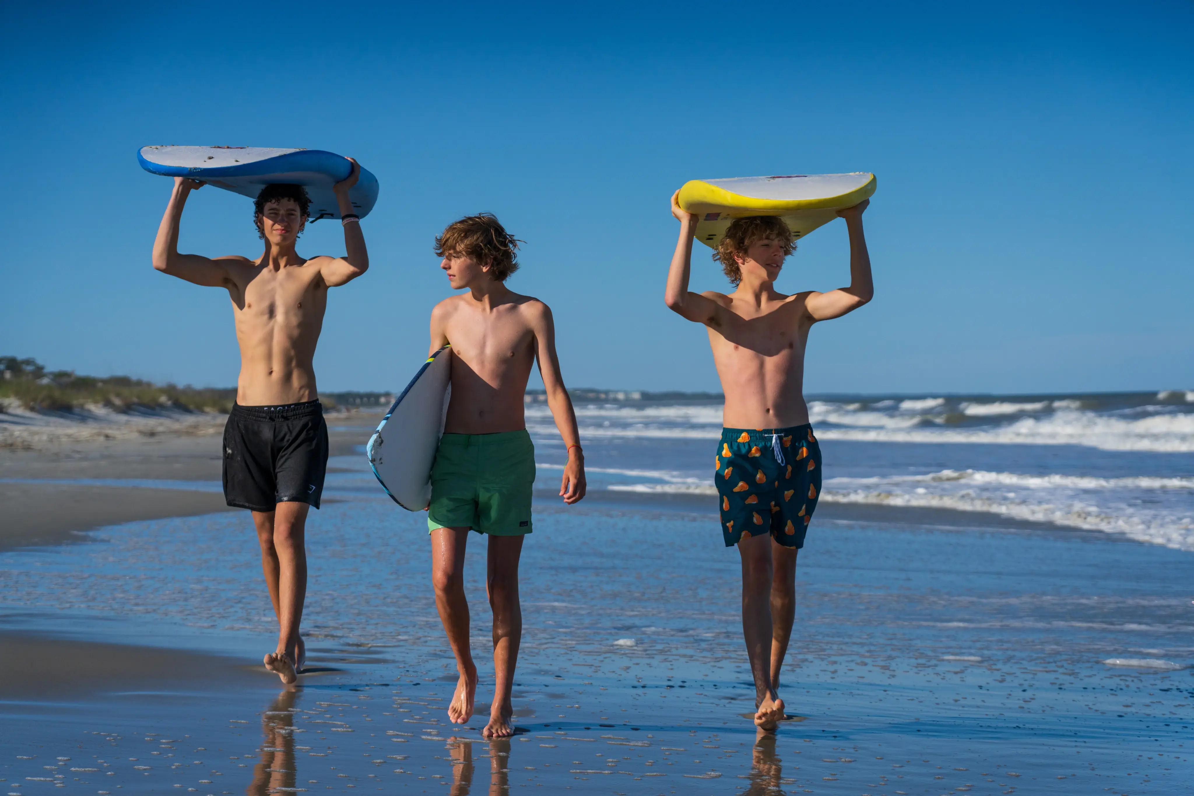 three young men carry surfboards over their heads and on their side as they walk the beach