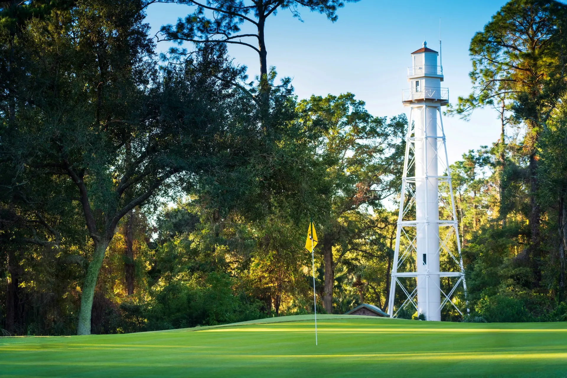 hole at Arthur Hills golf course with a yellow flag on the greens and white lighthouse  surrounded by trees in the background