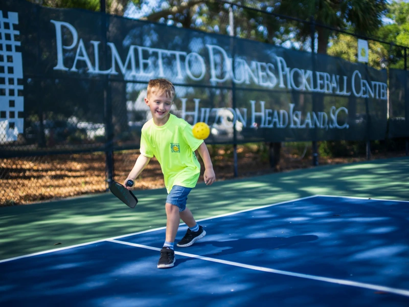 young boy in a bright yellow shirt actively playing pickleball at Palmetto Dunes Pickleball center