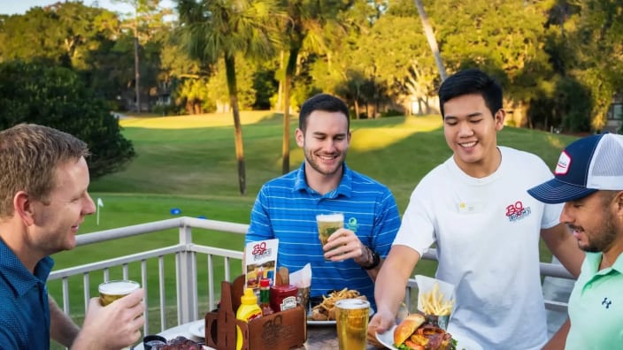 group of male golfers enjoy food and drinks on the Big Jim's deck overlooking the golf course while a Big Jim's server puts a plate of food down