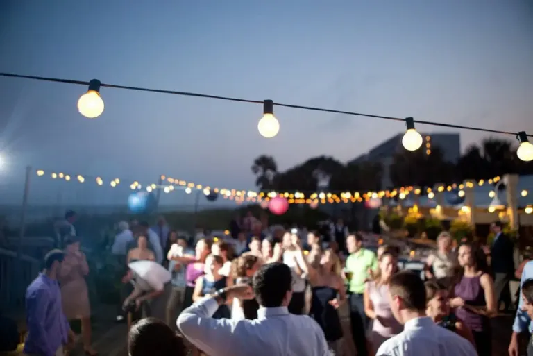 large group of wedding guests dance under bulb lights and the night sky on the open air deck