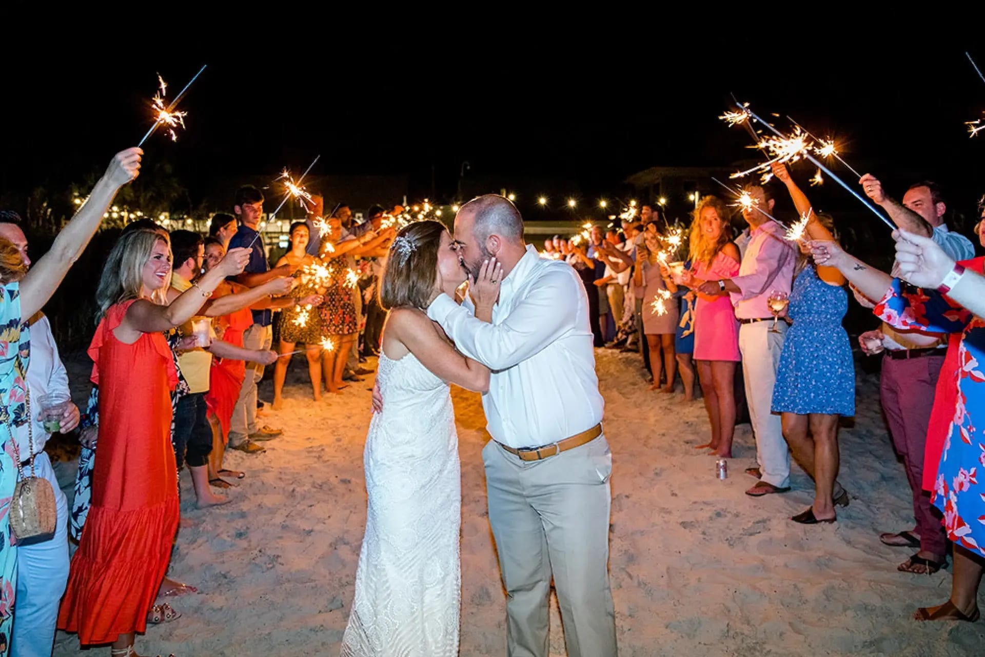 Newlyweds kissing on the beach at night, surrounded by guests with lit sparklers.