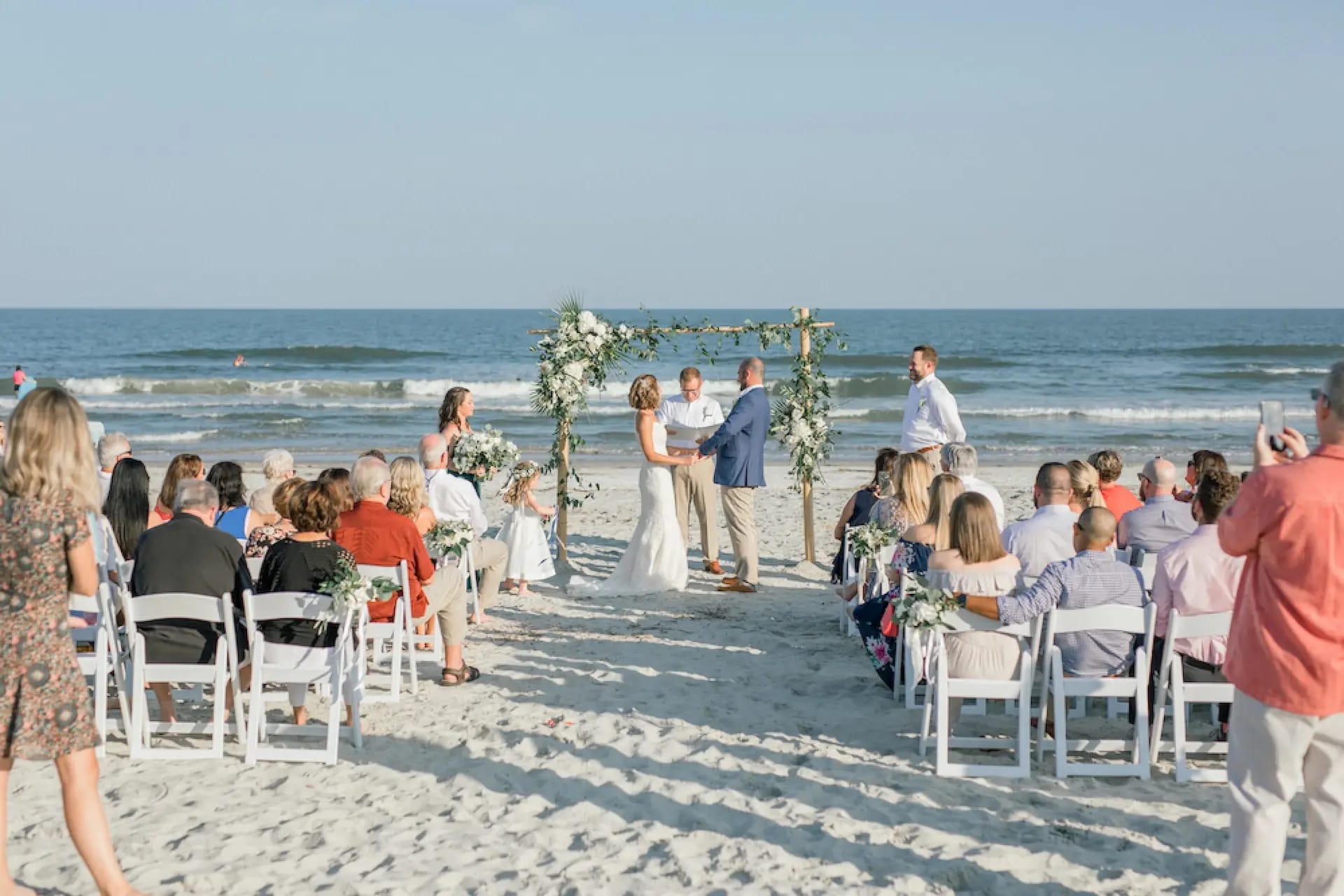 Beach wedding with the couple in front of the flower-decorated wooden altar and guests seated in white chairs.
