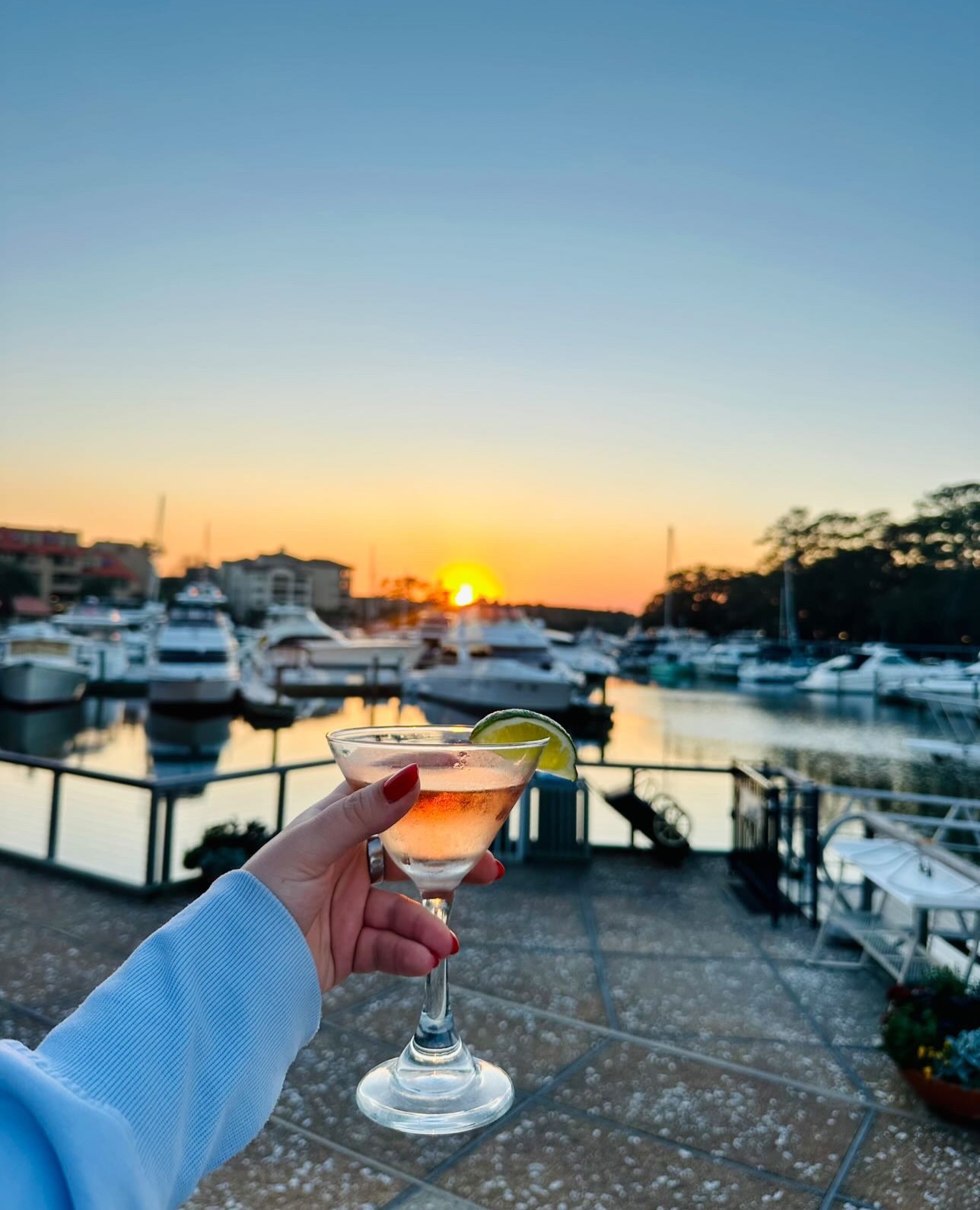 a womans hand with red nails holds up a cocktail in front of the marina with the sun setting in the background