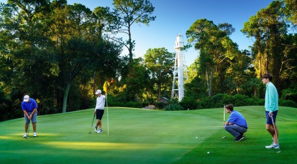 male golfers watch as a golfer putts on the golf course with the white lighthouse in the background