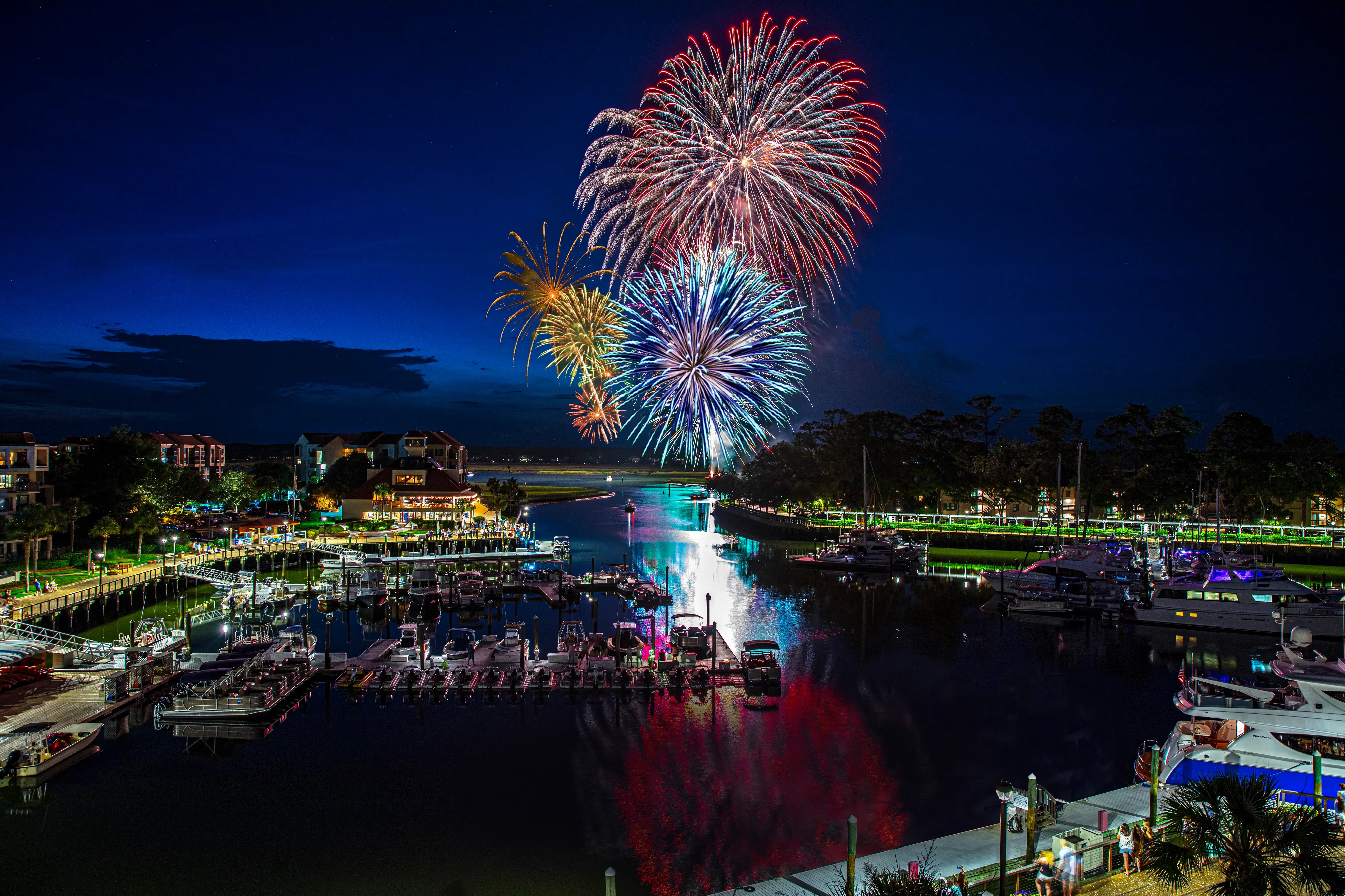 aerial view of the marina at night with boats and walkway lit up and fireworks exploding over the sky
