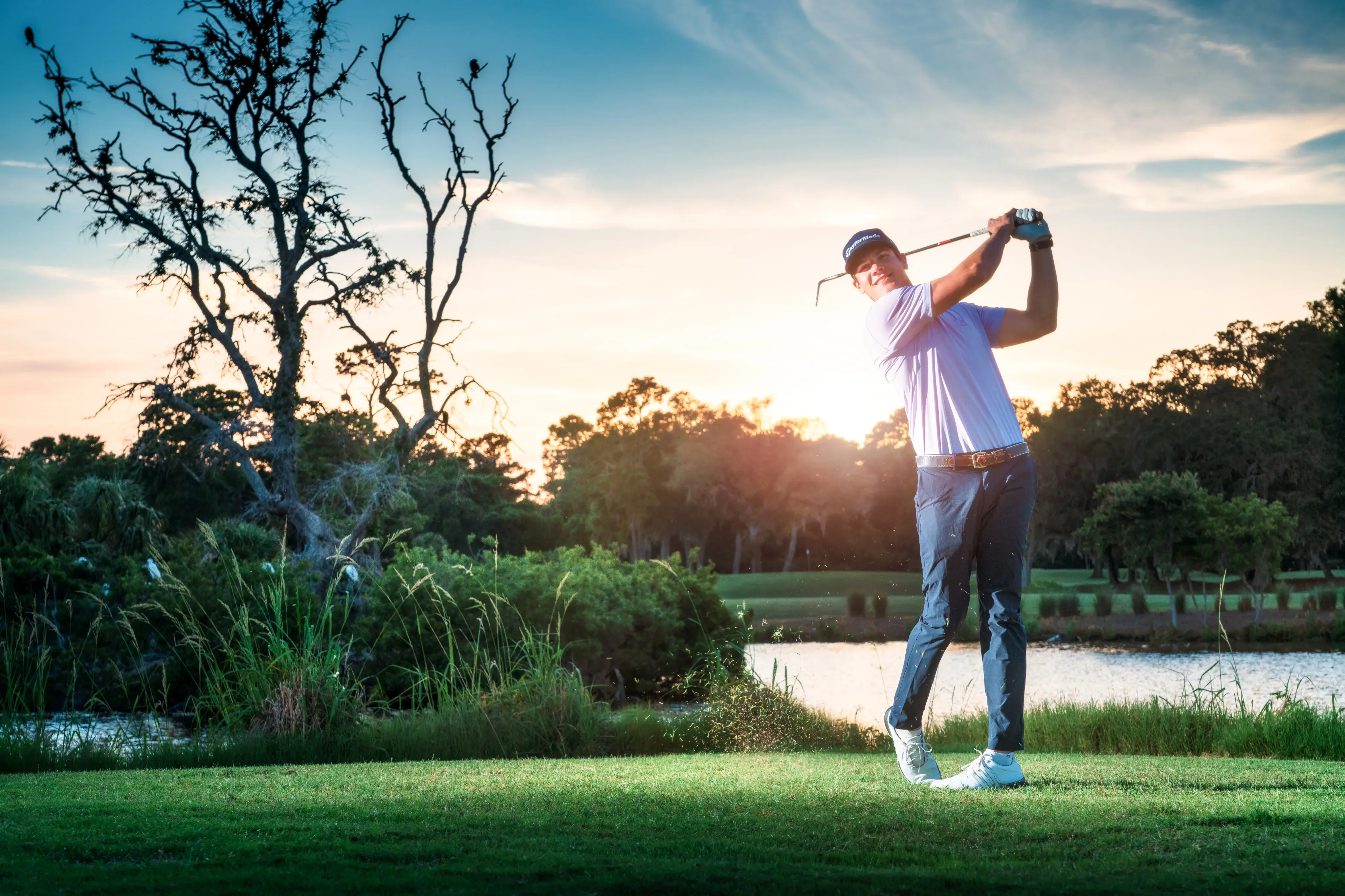 male golfer post swing on the golf course in front of water with the sun shining behind him