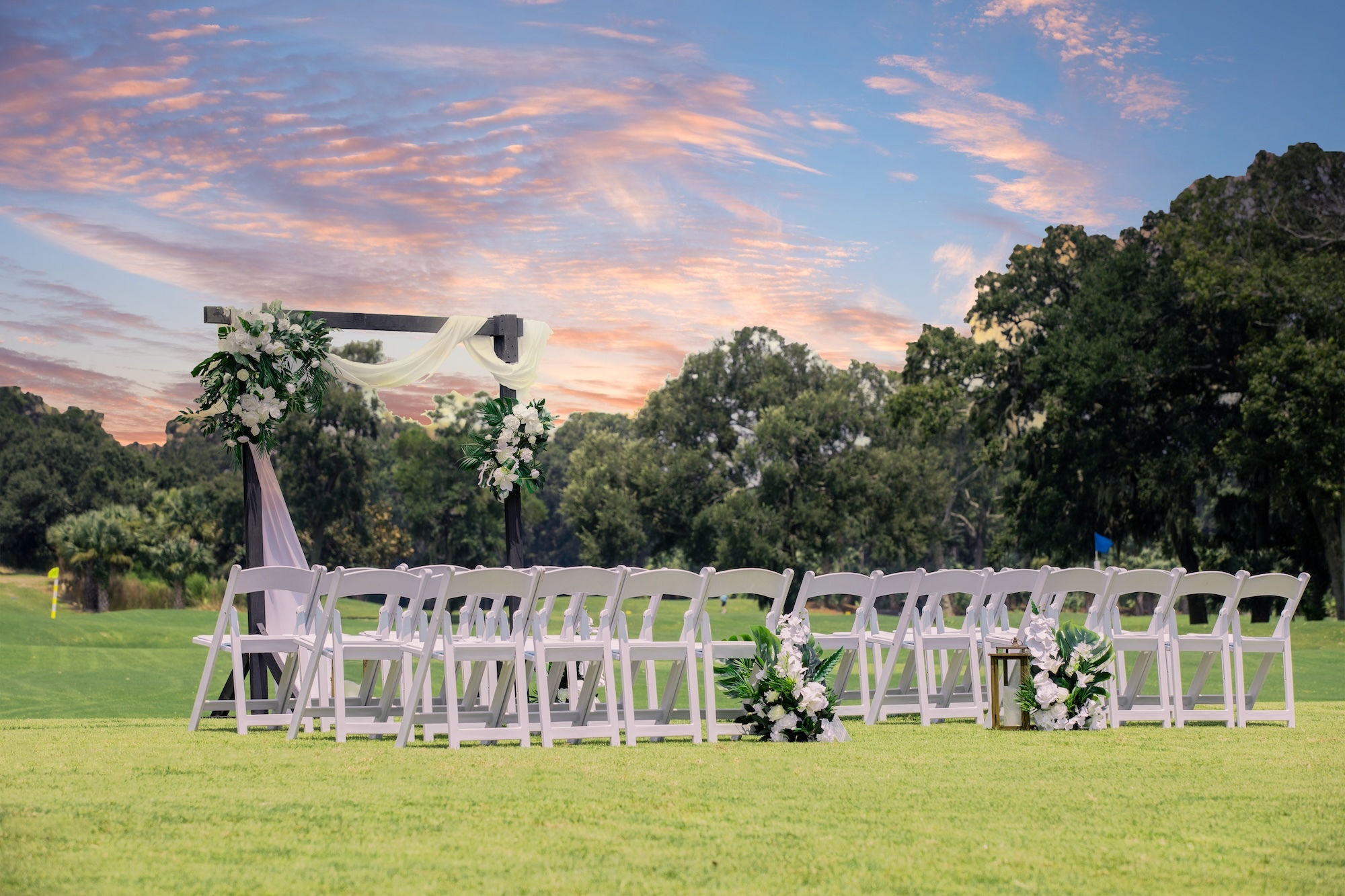 white chairs and a wooden arbor with flowers set up for a wedding ceremony on the grass under pastel skies