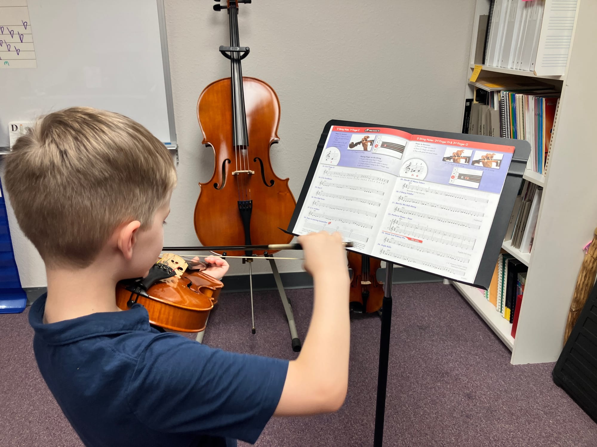Young violinist in a music lesson, holding a violin while reading sheet music on a stand, with a cello visible in the background.