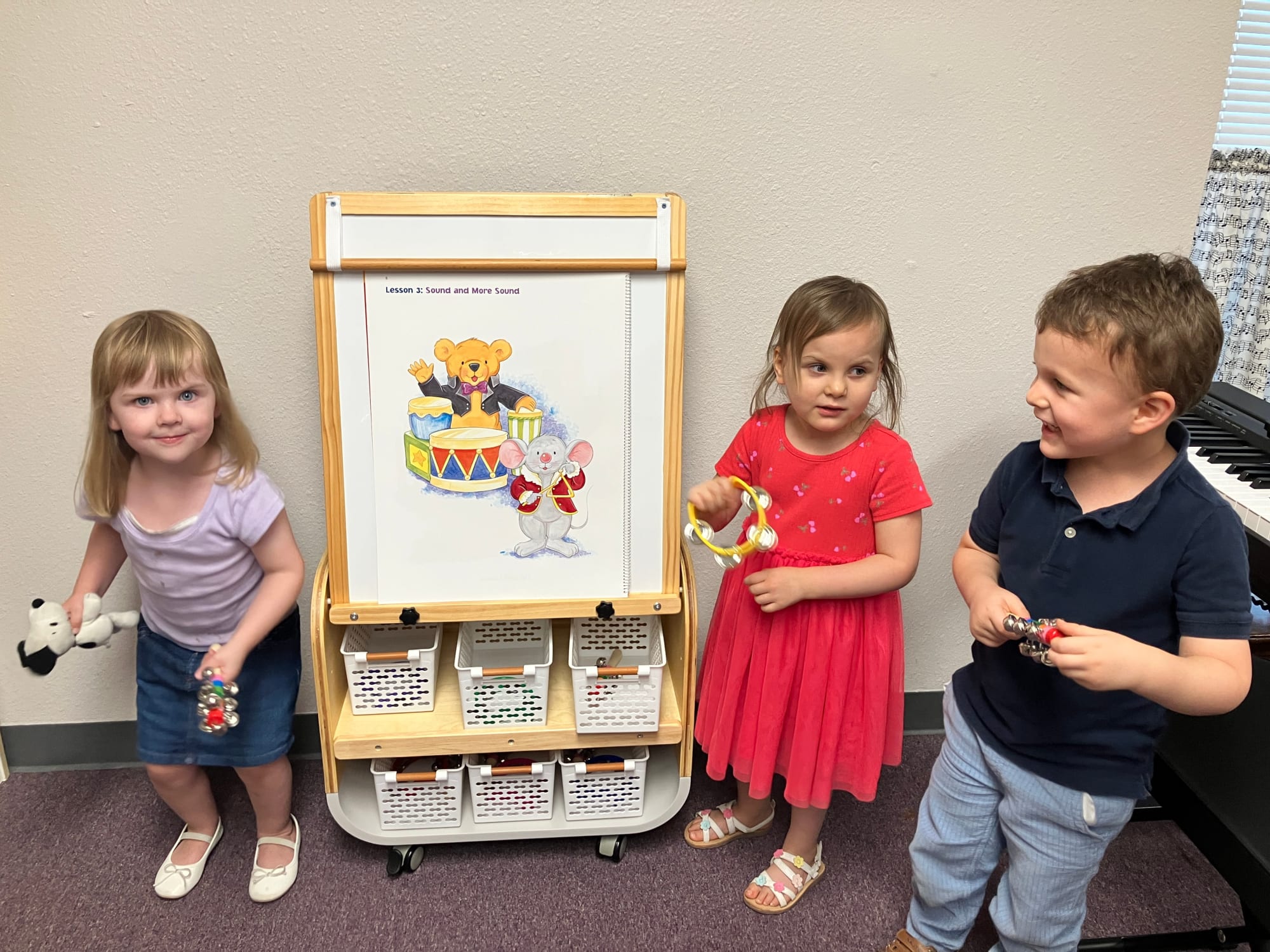 Four young children standing in a music lesson room next to a whiteboard displaying "Lesson 3: Sound and More Sound" with colorful illustrations of a