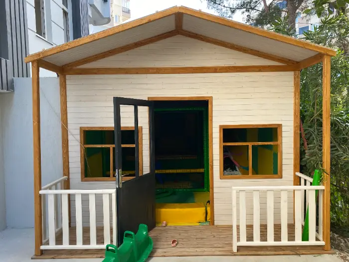 Yeşil ve sarı iç mekan oyun parkını barındıran, ahşap çatılı, beyaz panelli şirin bir dış cepheye sahip oyun evi.