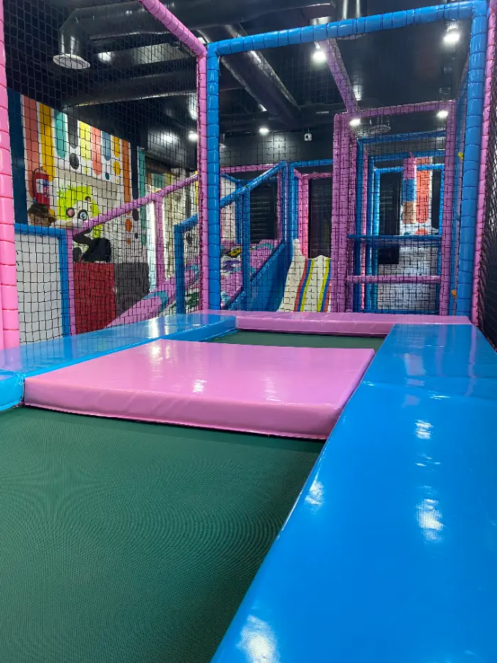 Çocukların güvenle zıplayabileceği, mavi ve pembe minderlerle çevrili, yeşil zeminli modern trambolin parkuru.