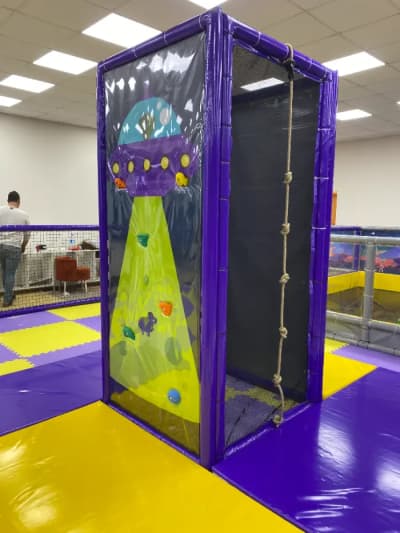 Purple and yellow soft play structure showing UFO-themed climbing wall and adjacent rope climbing section.