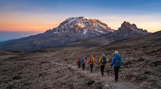 High altitude panorama of Mount Kilimanjaro snow capped summit Kibo illuminated by sunrise light, alpine desert landscape in foreground with high altitude hikers walking in single file line with trekking poles, crisp cool atmosphere.