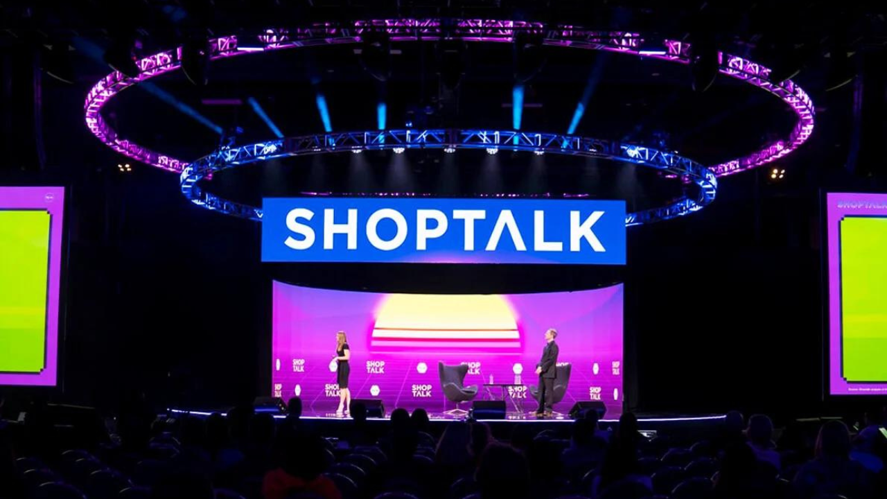 The image shows an interior view of a conference or event space during a presentation at the event "SHOPTALK". The stage is illuminated with a vibrant purple and blue lighting scheme. Above the stage, there's a large circular truss with lighting equipment, which is casting a dynamic glow on the surroundings. The backdrop of the stage prominently displays the event's name "SHOPTALK" in large white letters on a blue background. Two large vertical screens, one on each side of the stage, are lit up in bright neon colors. On the stage, there are two people, one standing on the far left and the other standing on the right. There are also several chairs set up on the stage, presumably for additional speakers or panelists. The audience area is dimly lit, with rows of empty seats visible in the foreground, suggesting that the event is either yet to start or currently in a break. The overall atmosphere of the image suggests a professional yet modern and tech-oriented event.