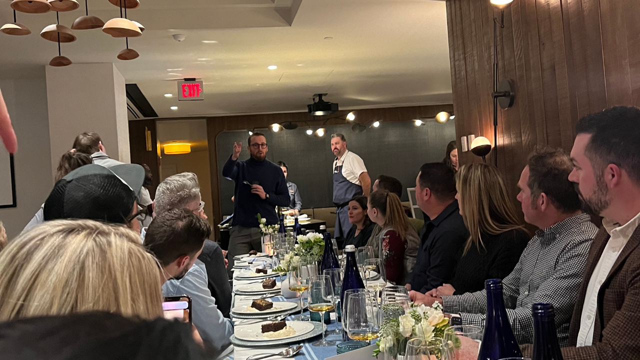 A scene from the SkiCommerce event dinner: attendees engage attentively as one person stands to speak, with elegant table settings and a warm, inviting atmosphere underscored by stylish décor.