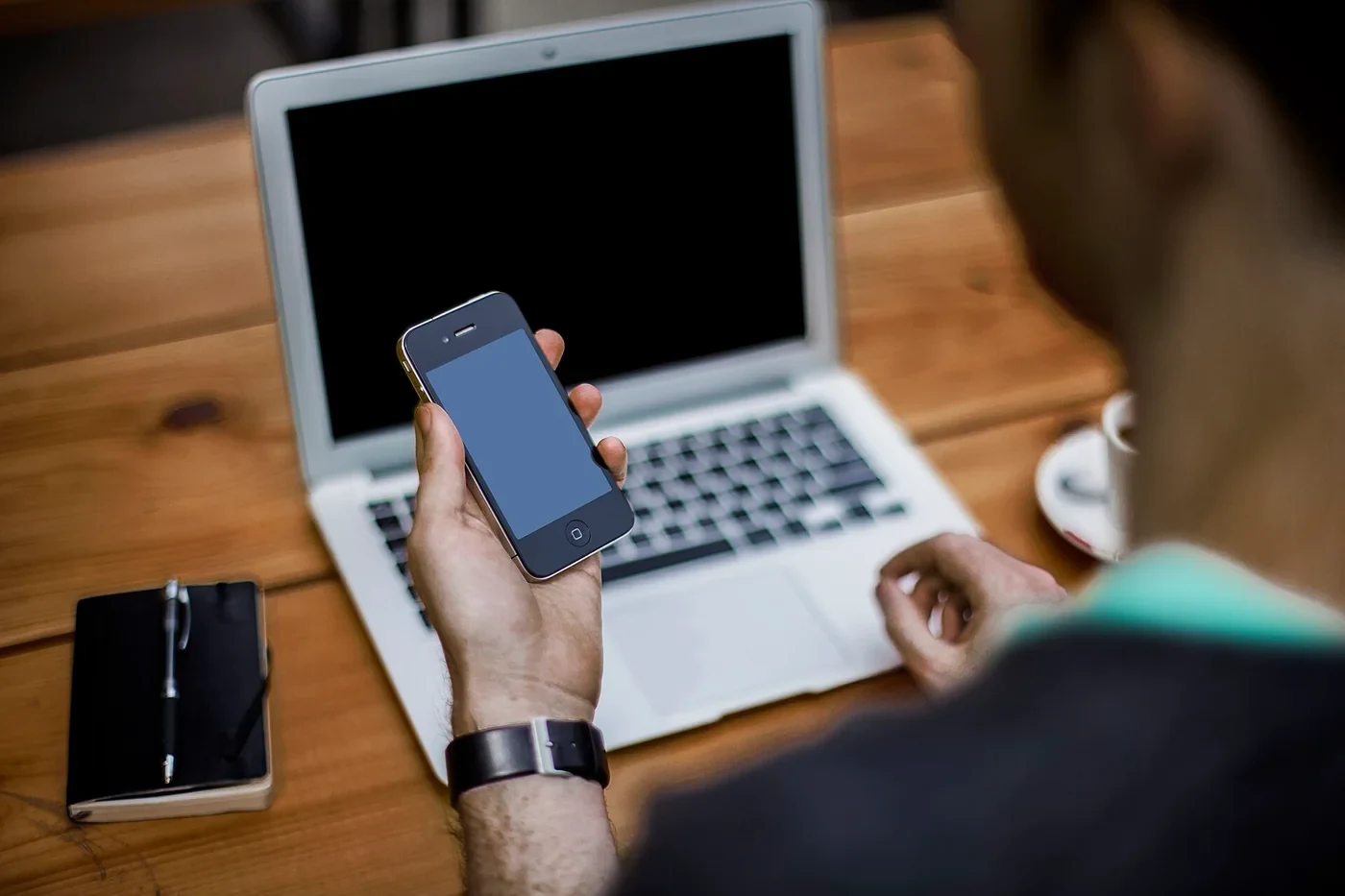 Une personne consulte son smartphone devant un ordinateur portable sur un bureau.