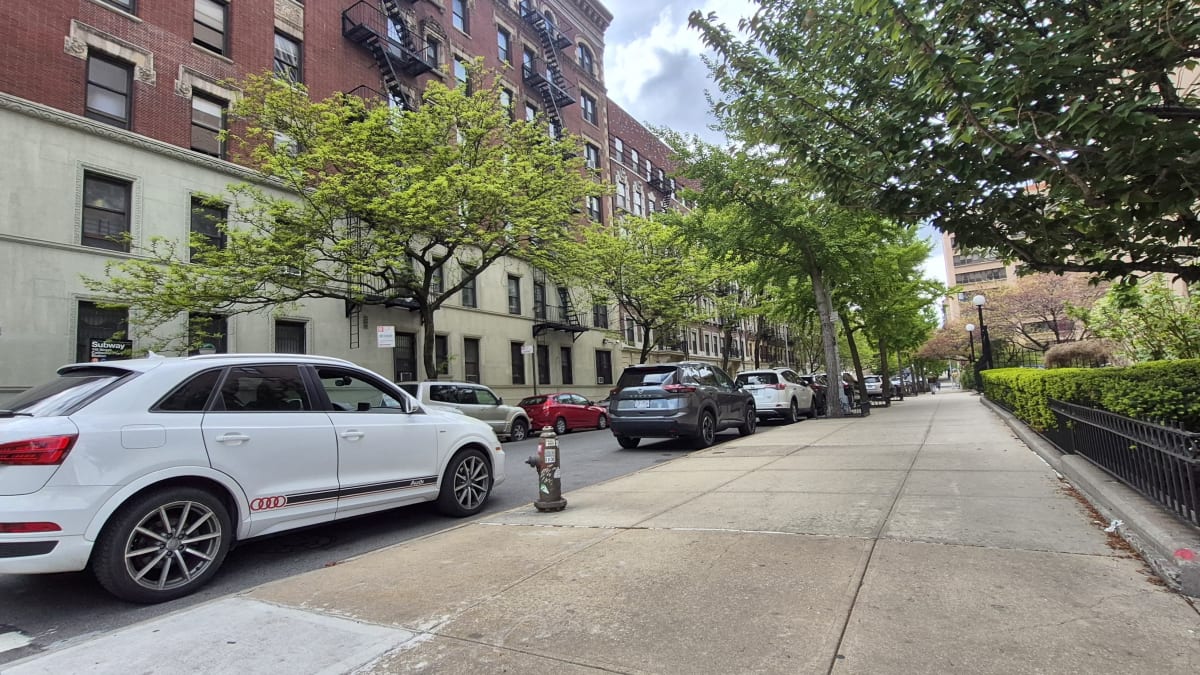 Red-brick prewar apartment buildings with fire escapes along a quiet residential street with parked cars