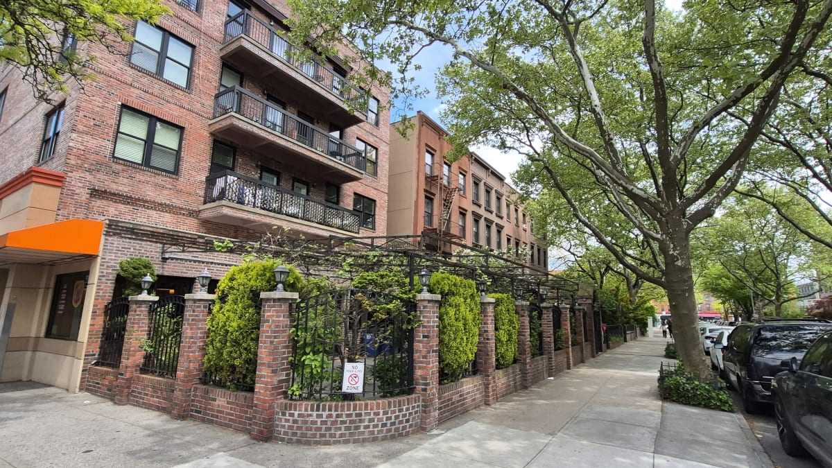 A red-brick apartment building with balconies and a hedged garden fence along a leafy residential sidewalk